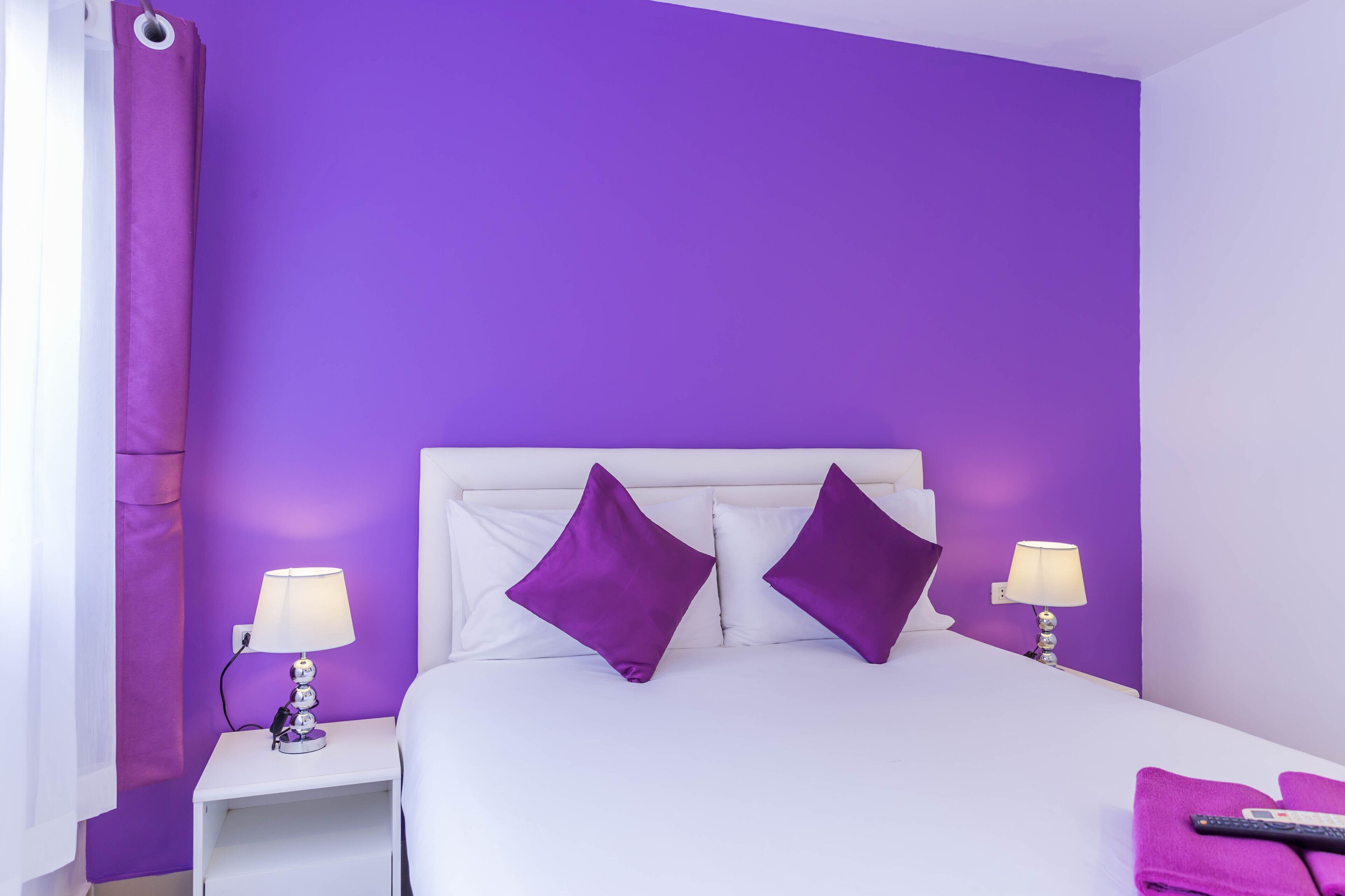 Een hotelkamer met een wit bed, paarse kussens en twee bedlampjes.