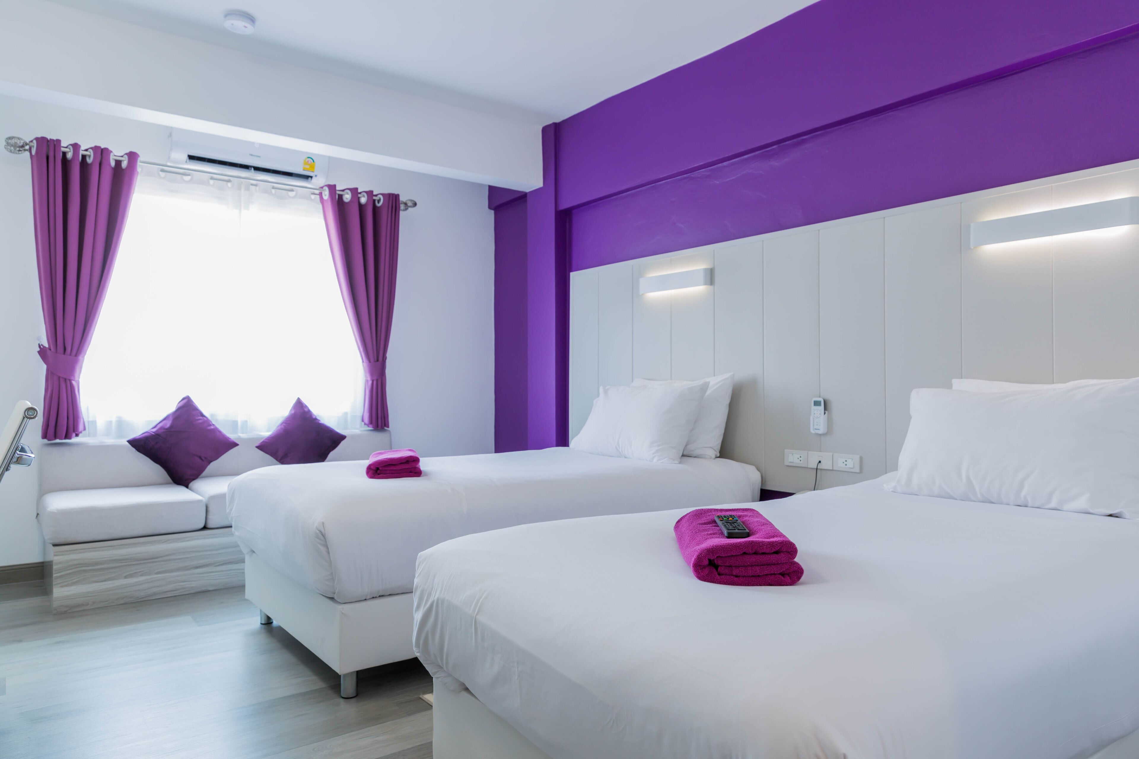 Habitación de hotel con dos camas, un sofá, cortinas moradas y una ventana con aire acondicionado.