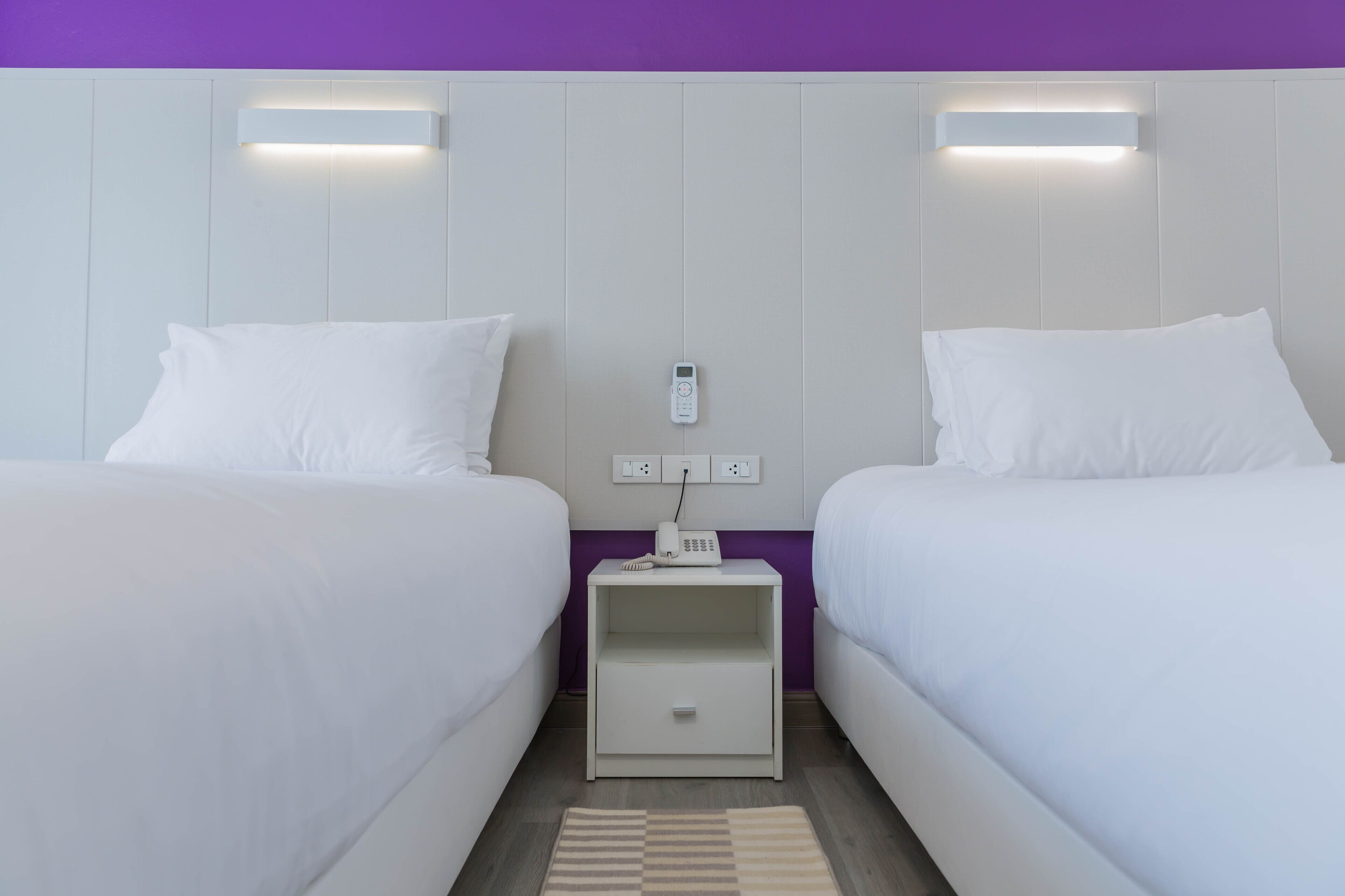 Habitación de hotel con dos camas, una mesita de noche con teléfono y toma de corriente, y una lámpara fijada a la pared.