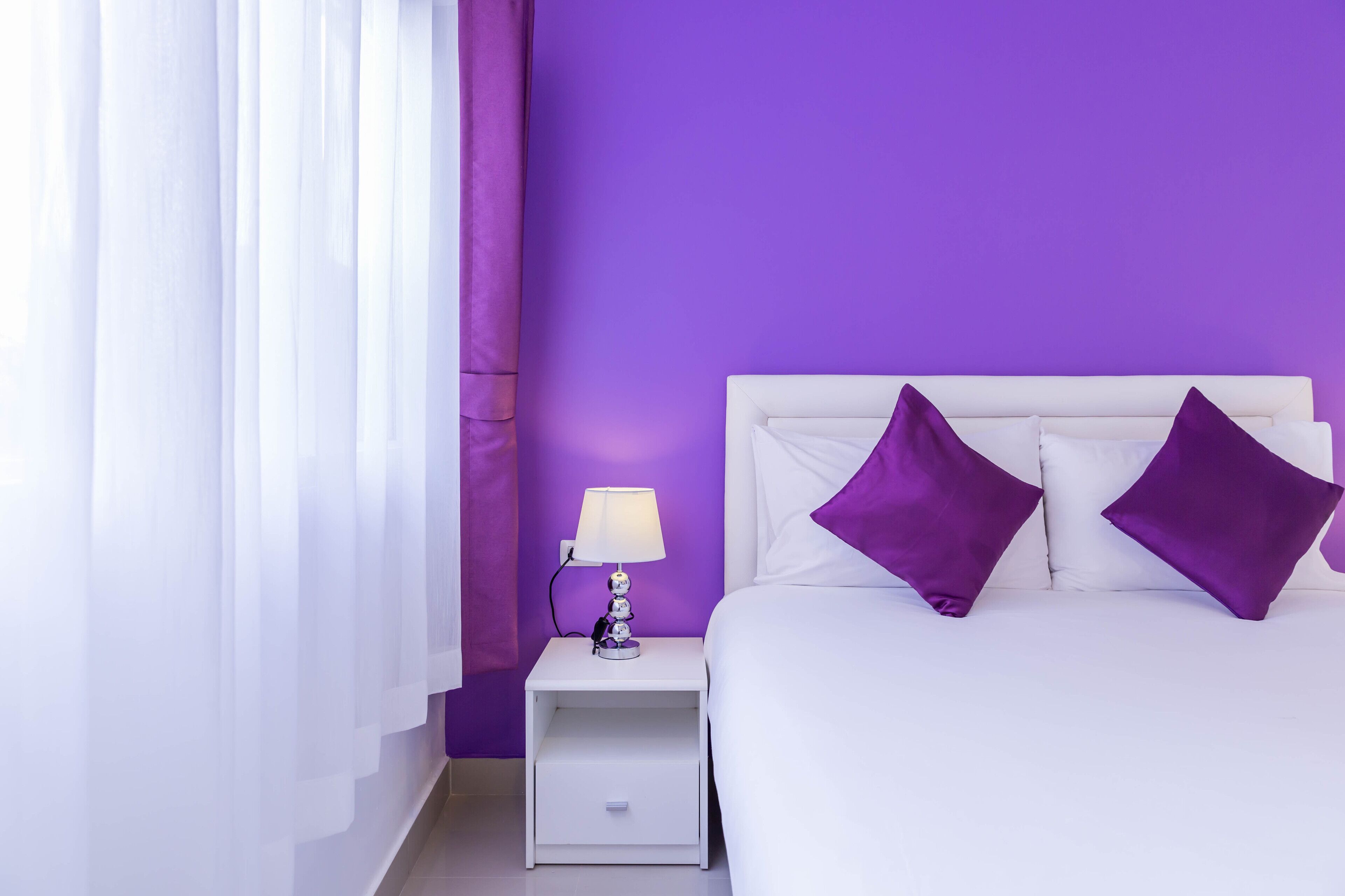Ein Schlafzimmer mit einem weißen Bett, violetten Kissen, einem weißen Nachttisch mit einer Lampe und einer violetten Wand.