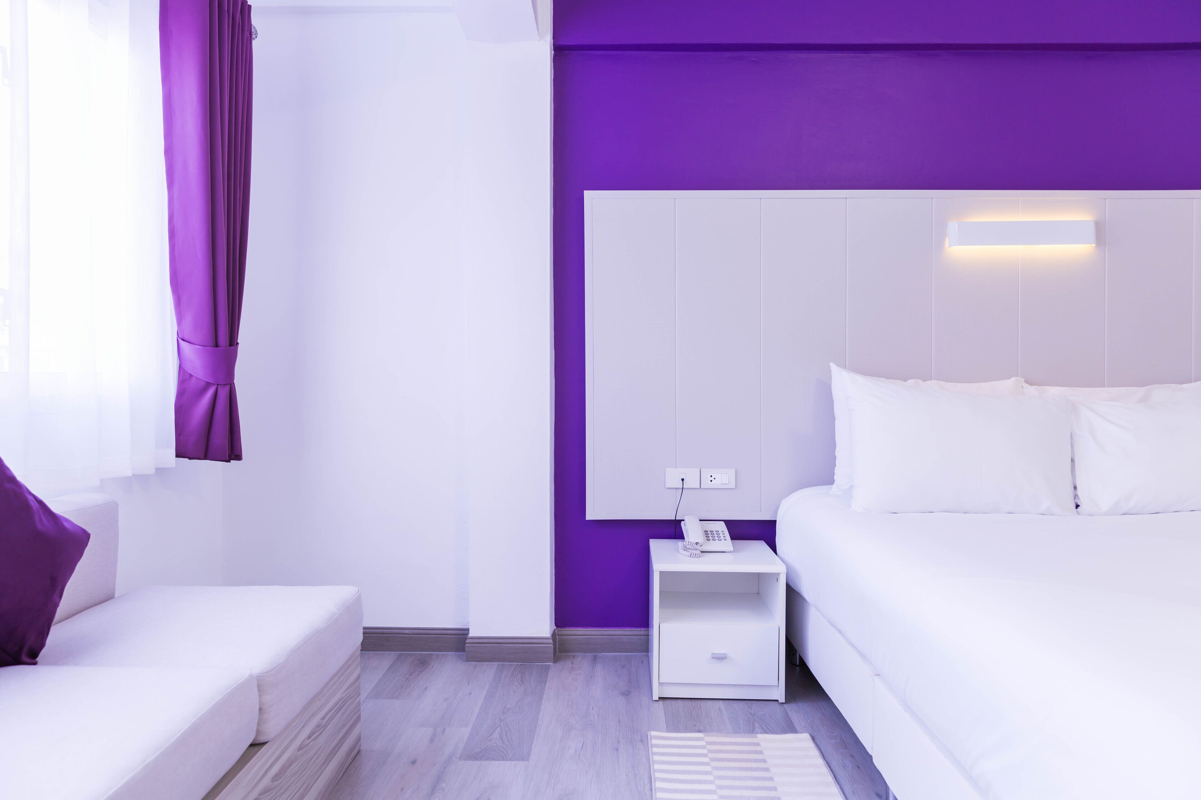 A modern hotel room with a white bed, a nightstand with a phone, and a purple accent wall.