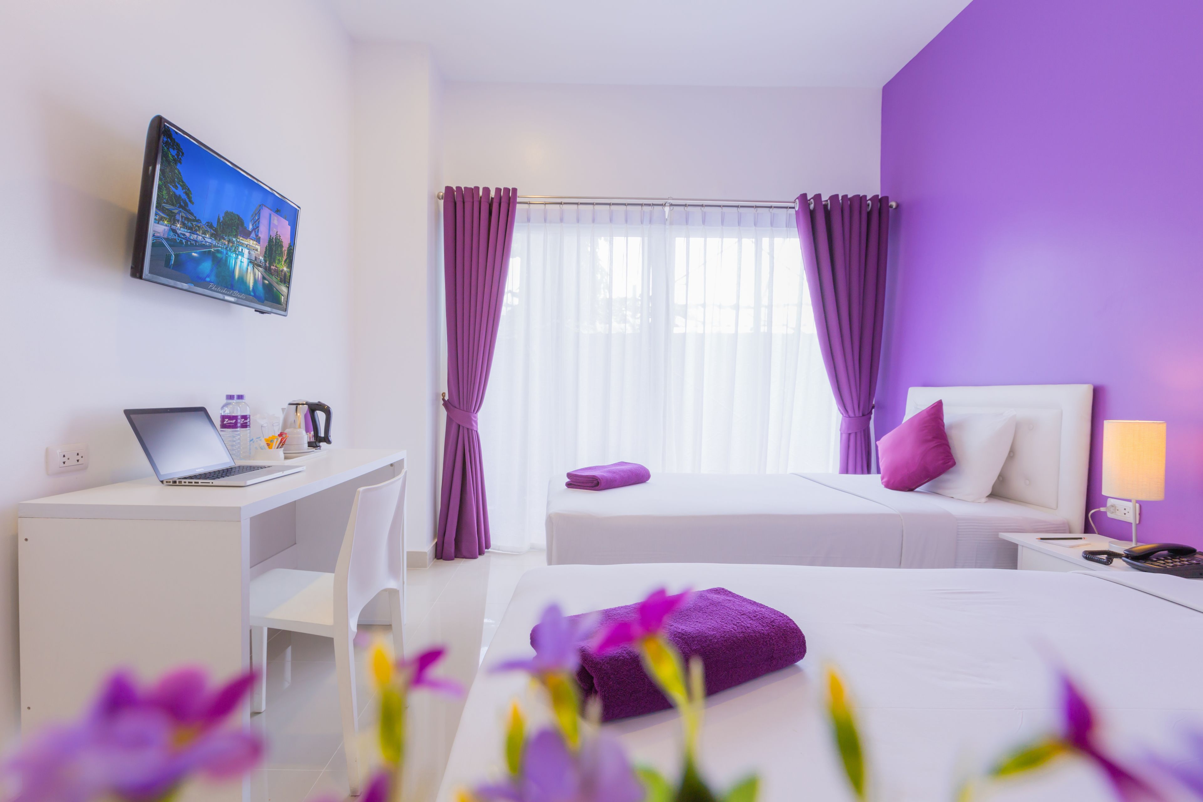 A hotel room with a bed, a desk with a laptop, a chair, a nightstand, and a window with curtains.