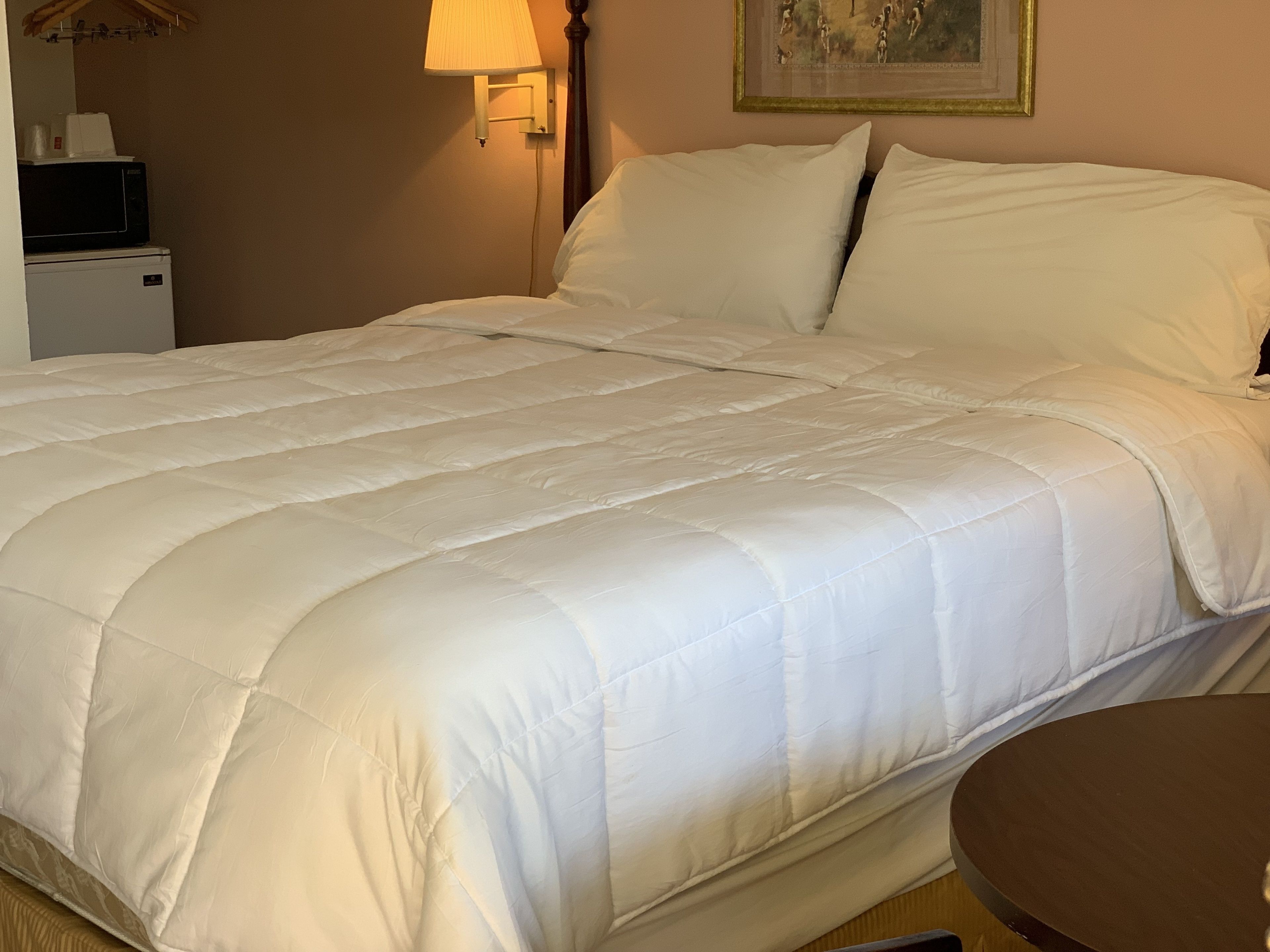A neatly made bed with a white duvet, a bedside table with a lamp, and a small refrigerator.