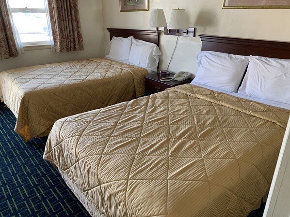 Dos camas tamaño queen con colchas color beige, una mesita de noche con teléfono y una ventana con cortinas.