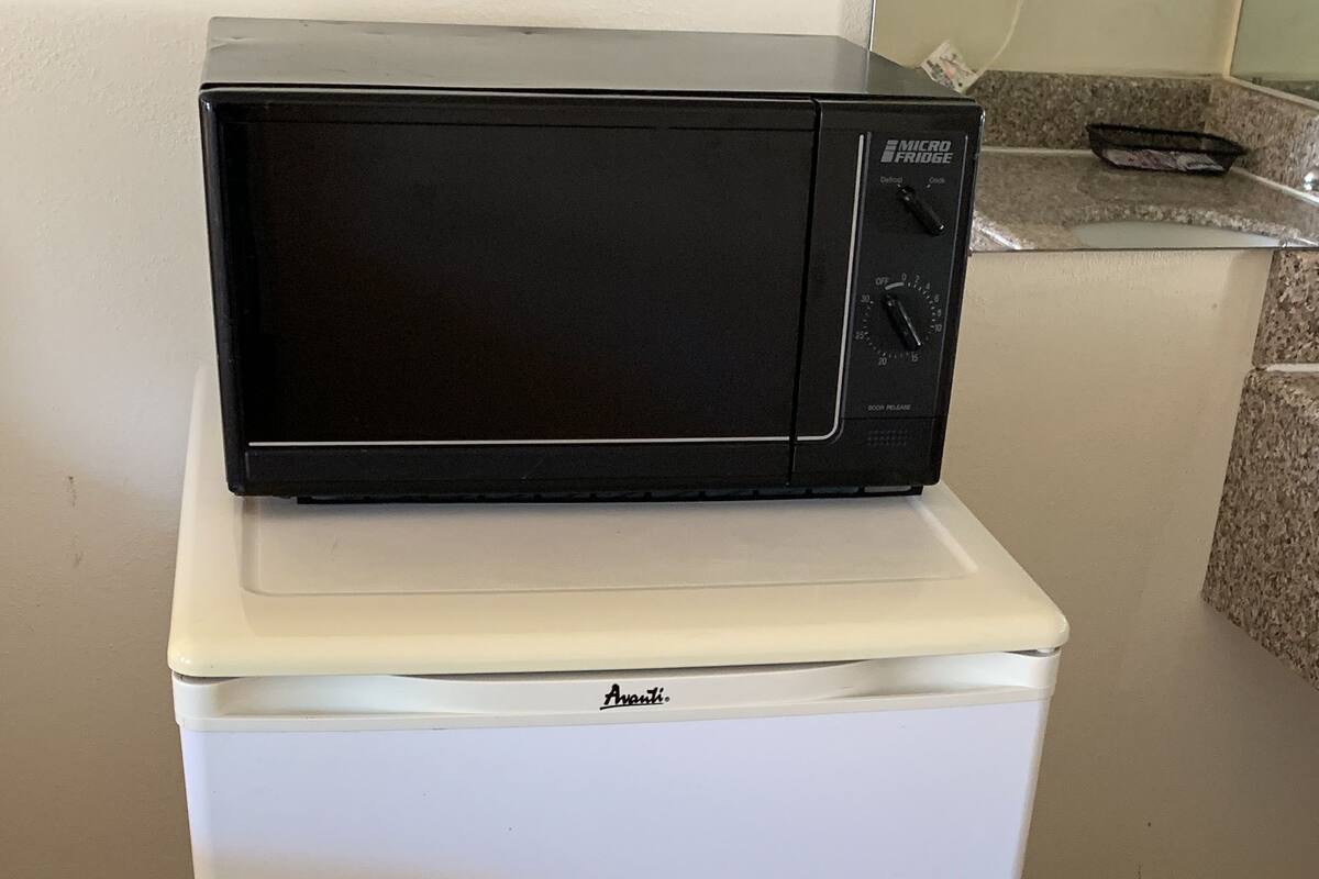 A white mini-fridge with a black microwave on top, a power strip, and a trash can in the corner.