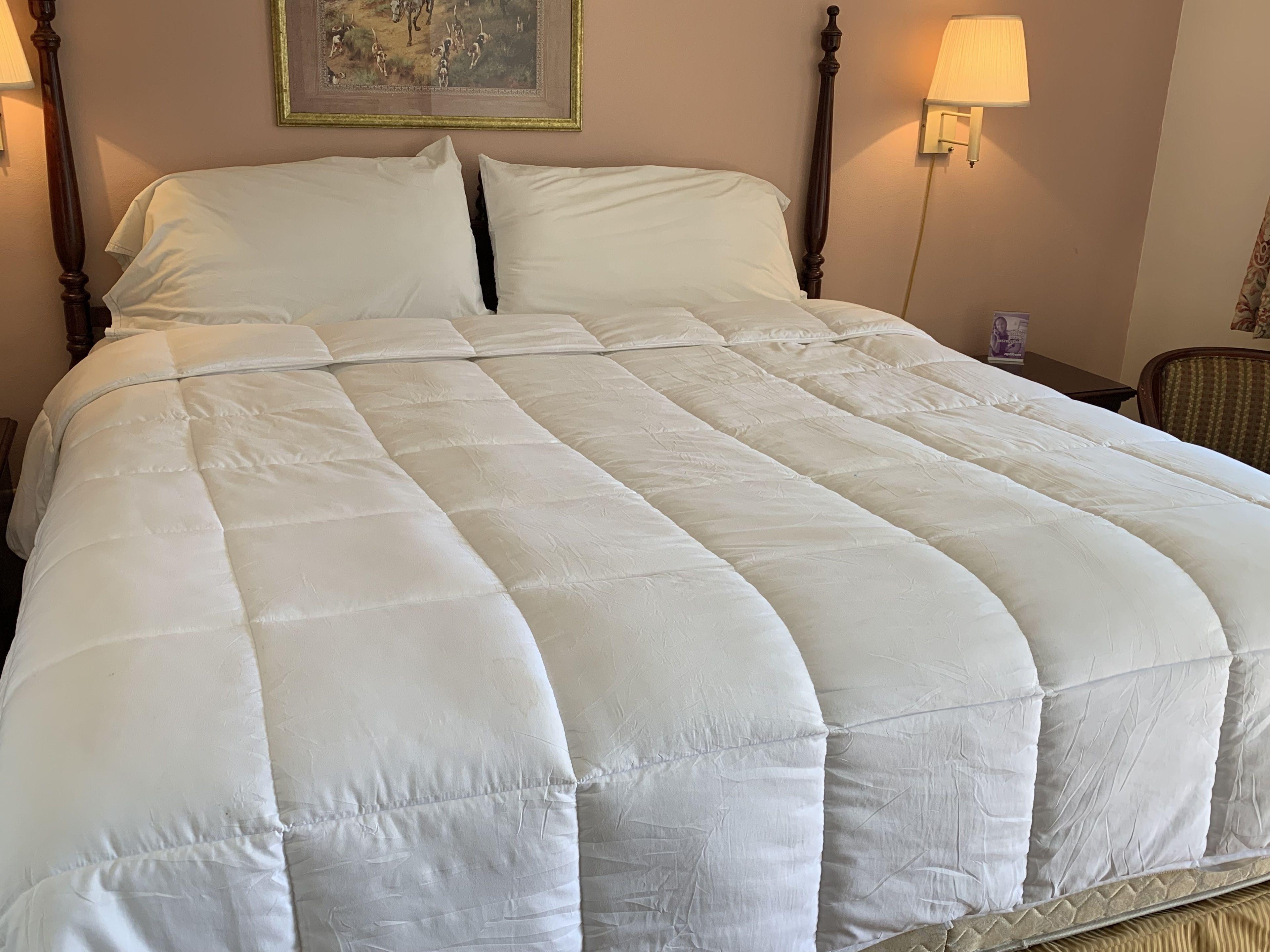A large bed with a white quilted bedspread, two bedside lamps, and a framed painting on the wall.
