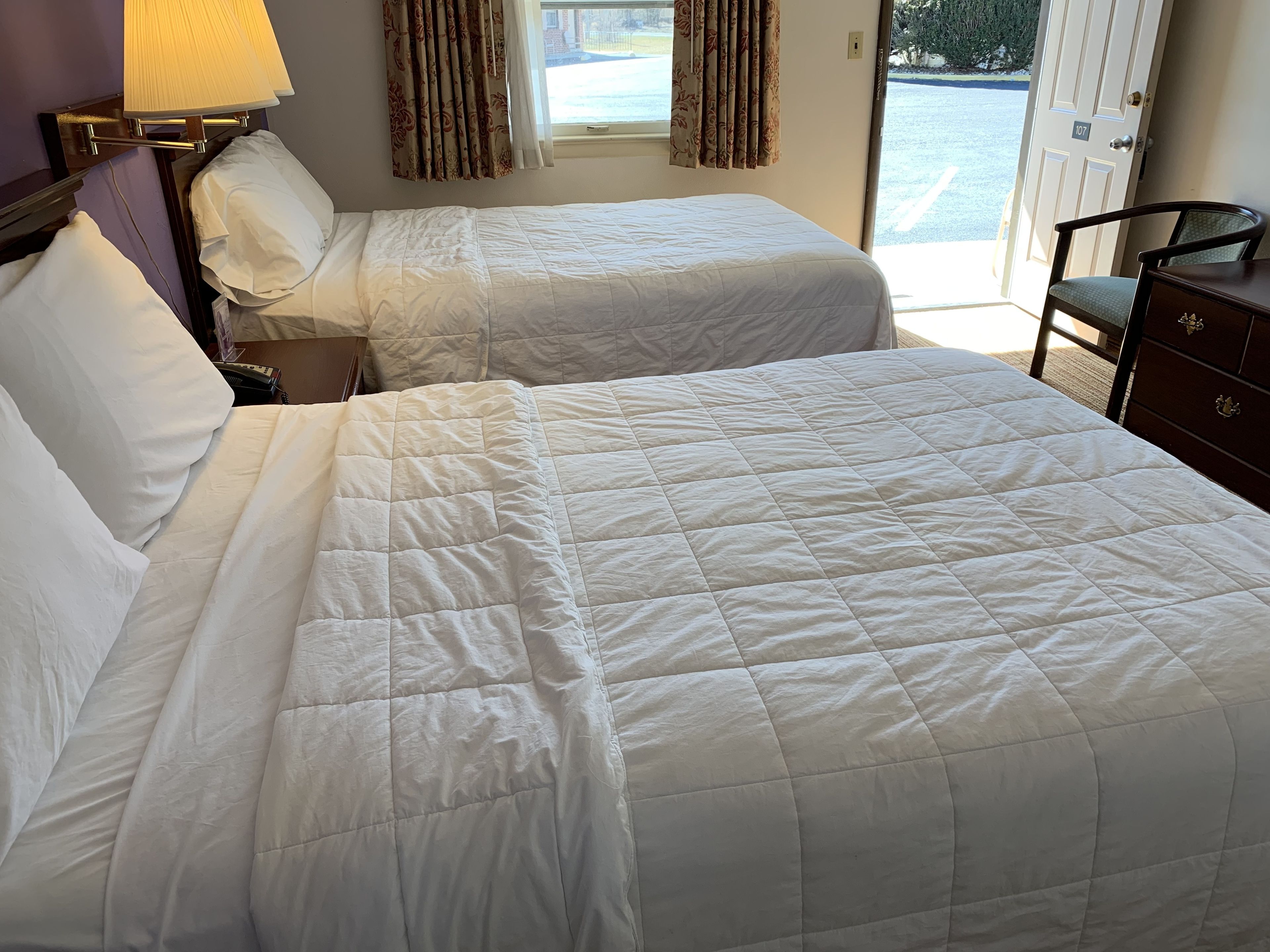 A hotel room with two beds, a nightstand, a lamp, a dresser, and a view of the outside.