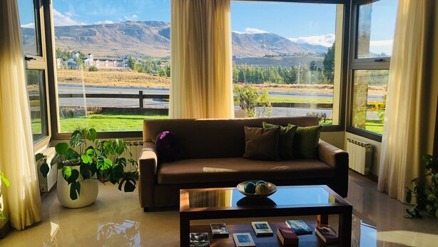Uma sala de estar moderna com um sofá, uma mesa de centro com livros e janelas amplas com vista para uma paisagem montanhosa.