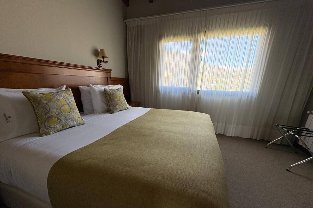 Une chambre d’hôtel avec un grand lit, une tête de lit en bois, une fenêtre avec des rideaux blancs et une petite table avec une lampe.