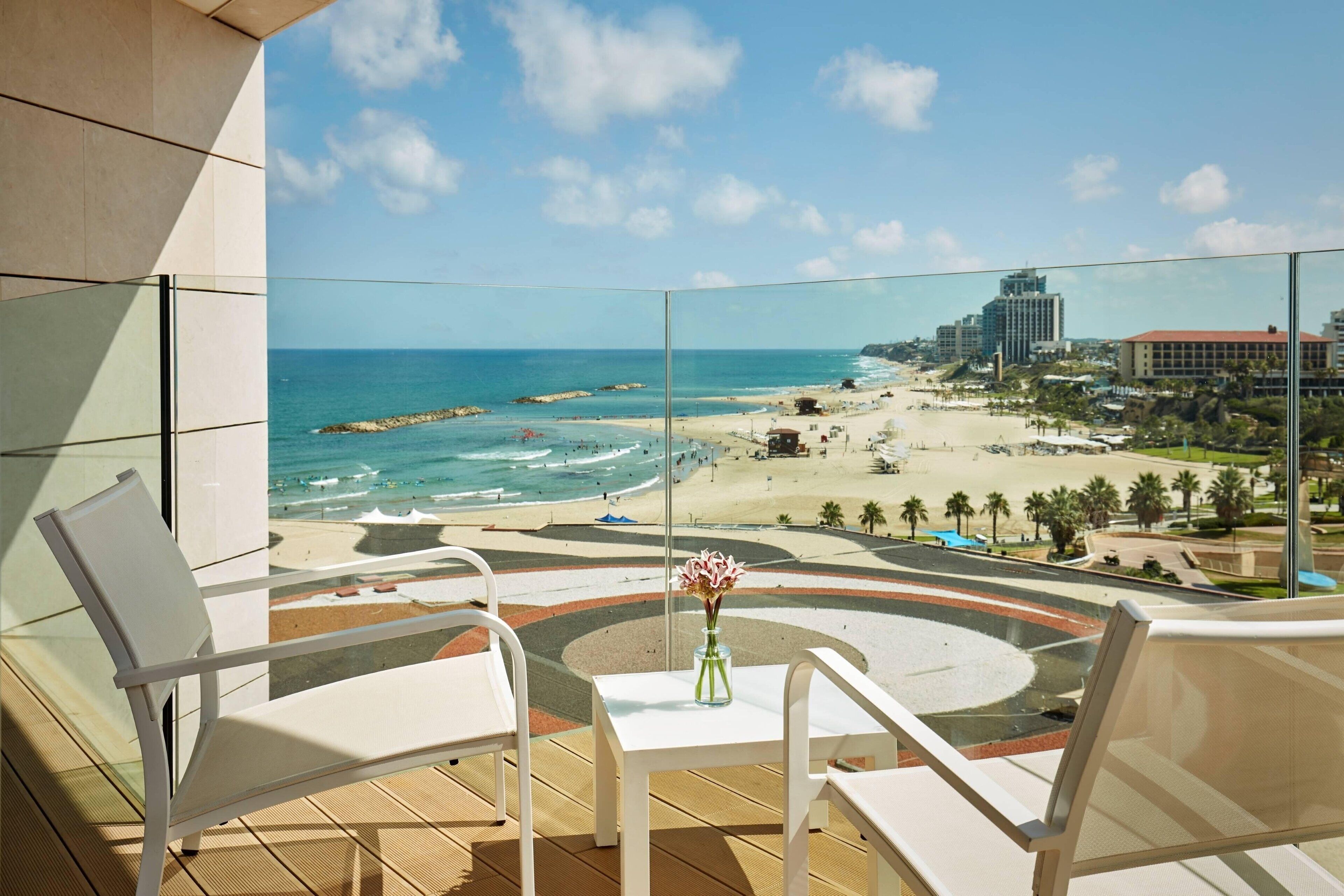 Un balcon doté d’une balustrade en verre, offrant une vue sur une plage et l’océan. On y trouve des chaises blanches et une petite table sur laquelle est posé un vase de fleurs.