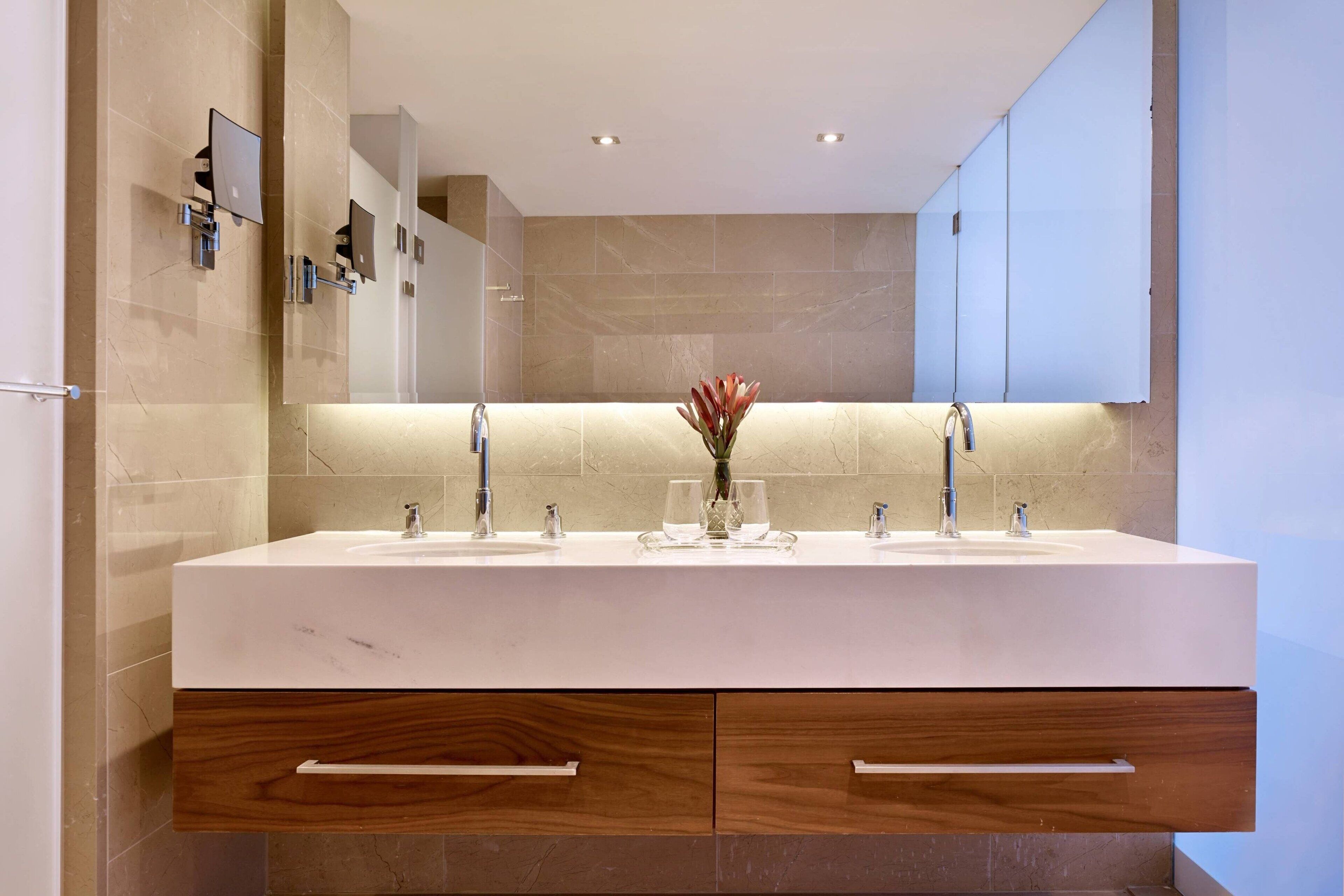 A modern bathroom with a large white sink, two faucets, and a mirror above. There are wooden cabinets below the sink and a vase with flowers on the counter.