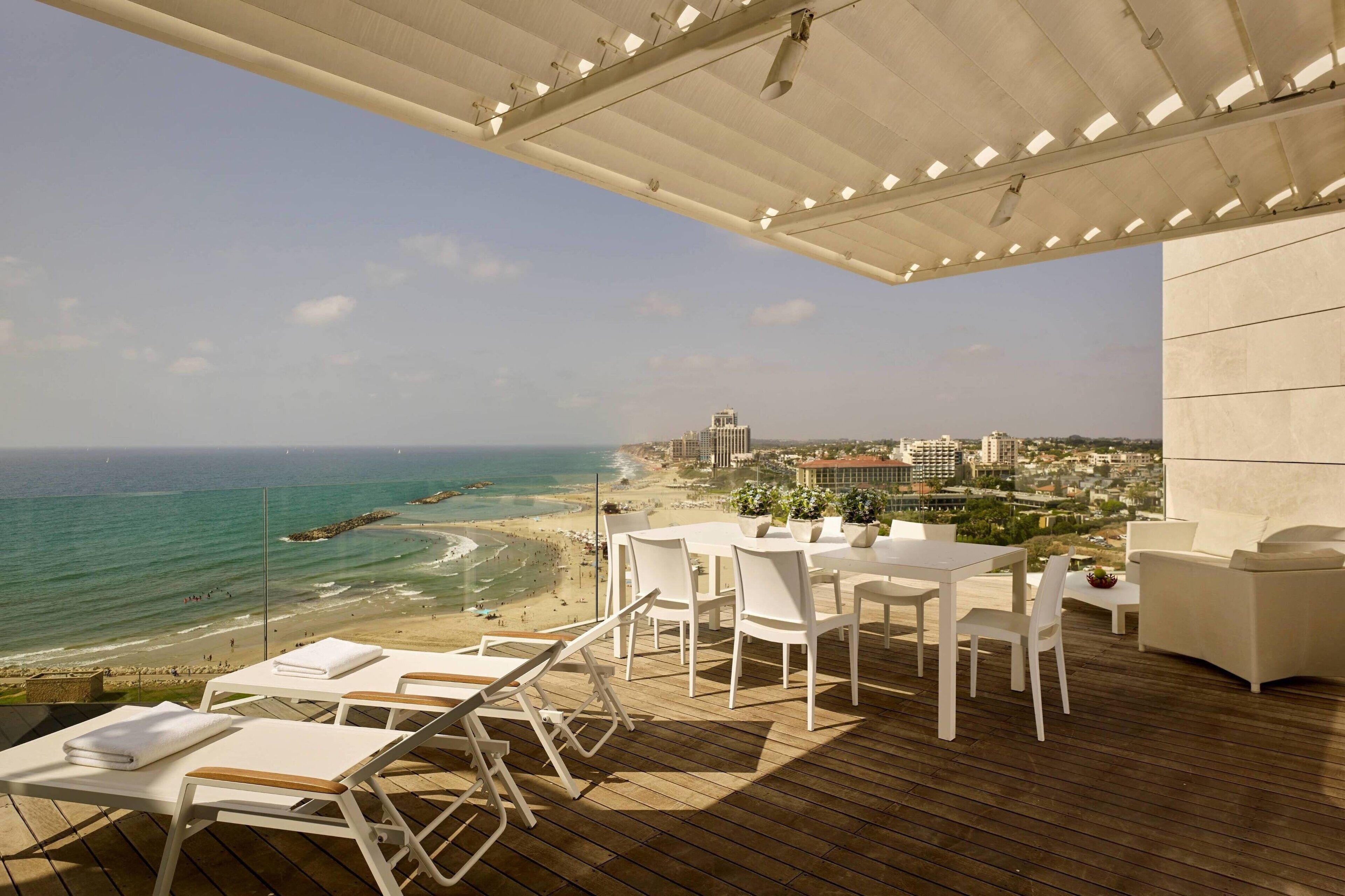 Terraço com vista para a praia e oceano, com mobiliário branco e plantas em vasos.