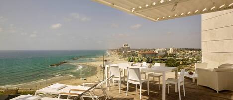 Terraço com vista para a praia e oceano, com mobiliário branco e plantas em vasos.