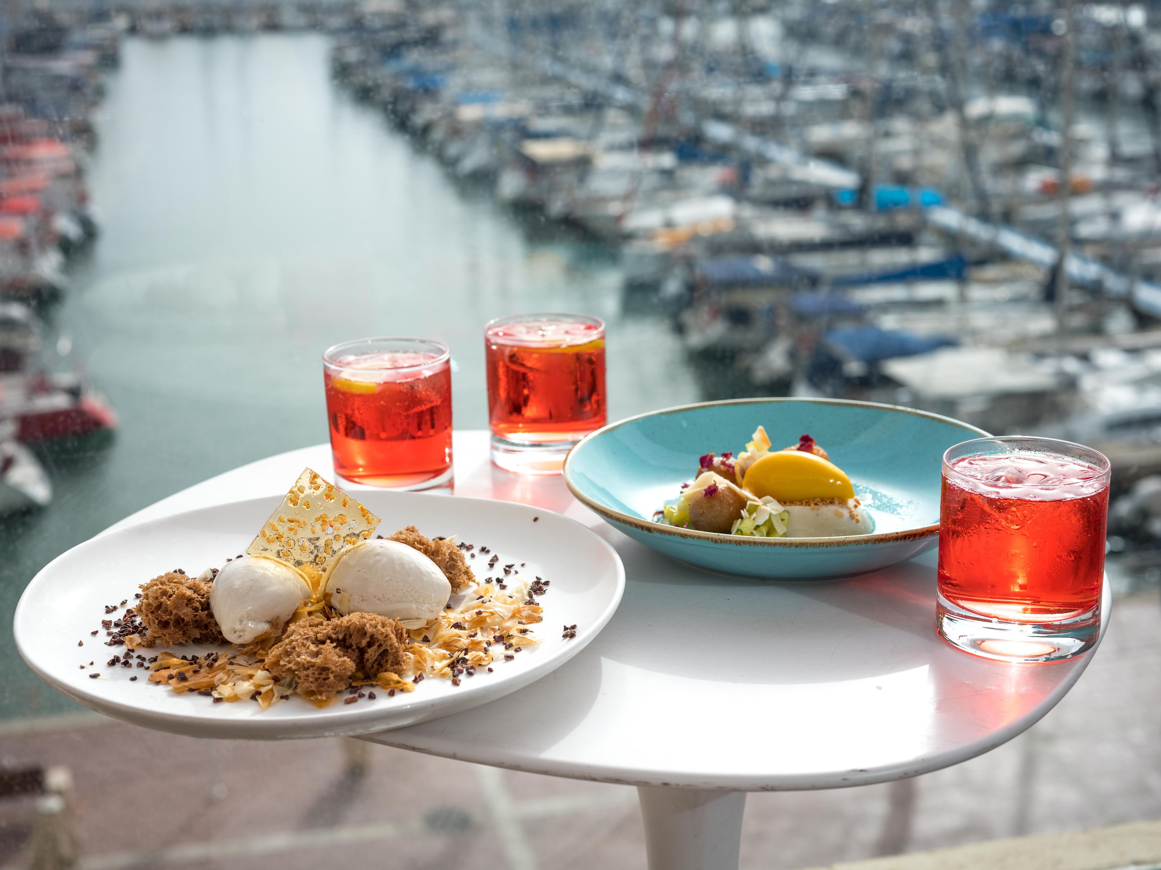 Et bord med en desserttallerken, to glas med rød drik og en blå skål med mad, med udsigt over en havn med både.