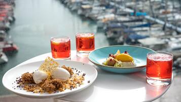 Una mesa con un plato de postre, dos vasos de bebida roja y un bol azul con comida, con vista a un puerto deportivo lleno de barcos.