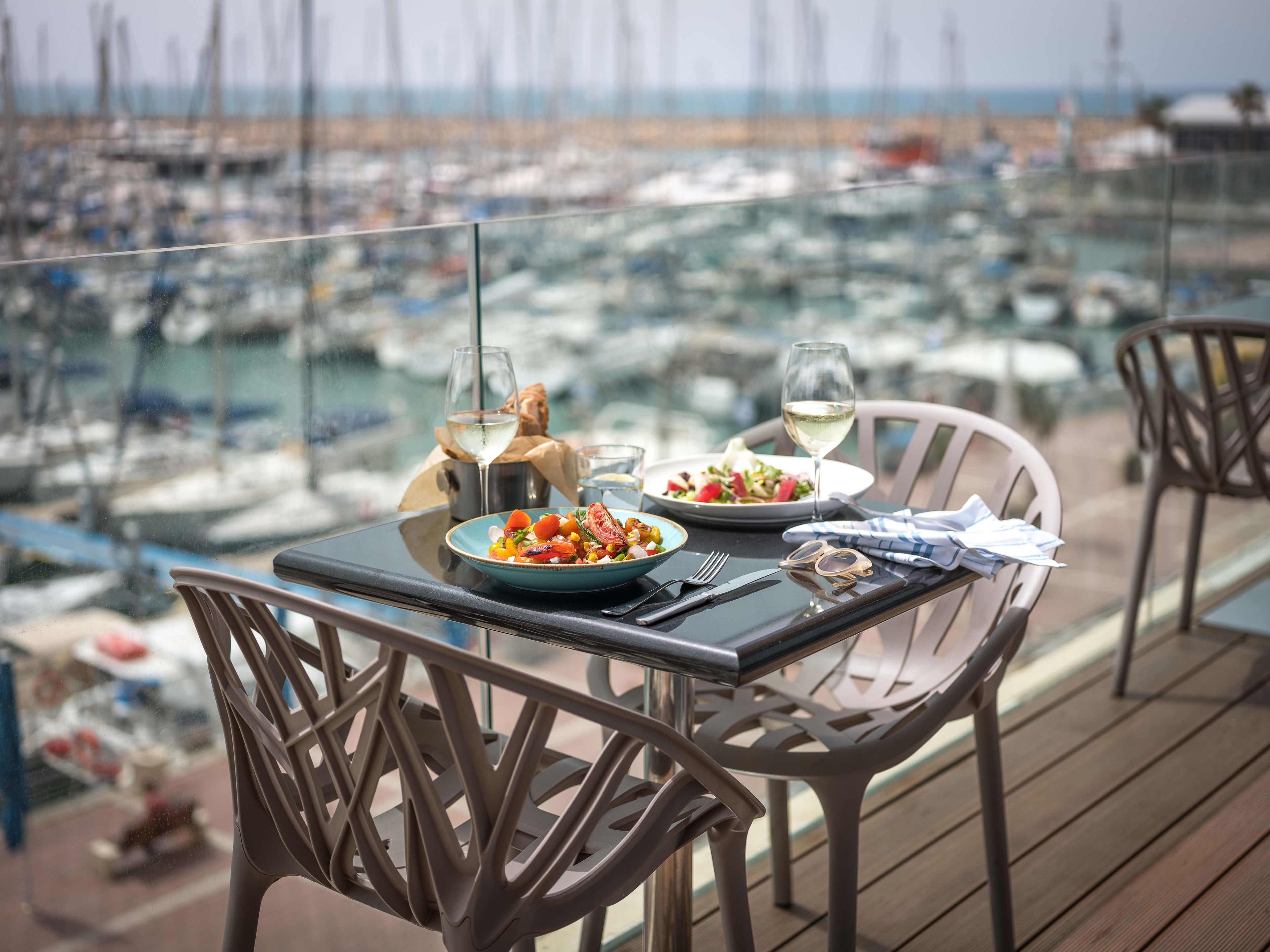 A dining table with plates of food, wine glasses, and cutlery on a balcony overlooking a marina with boats.