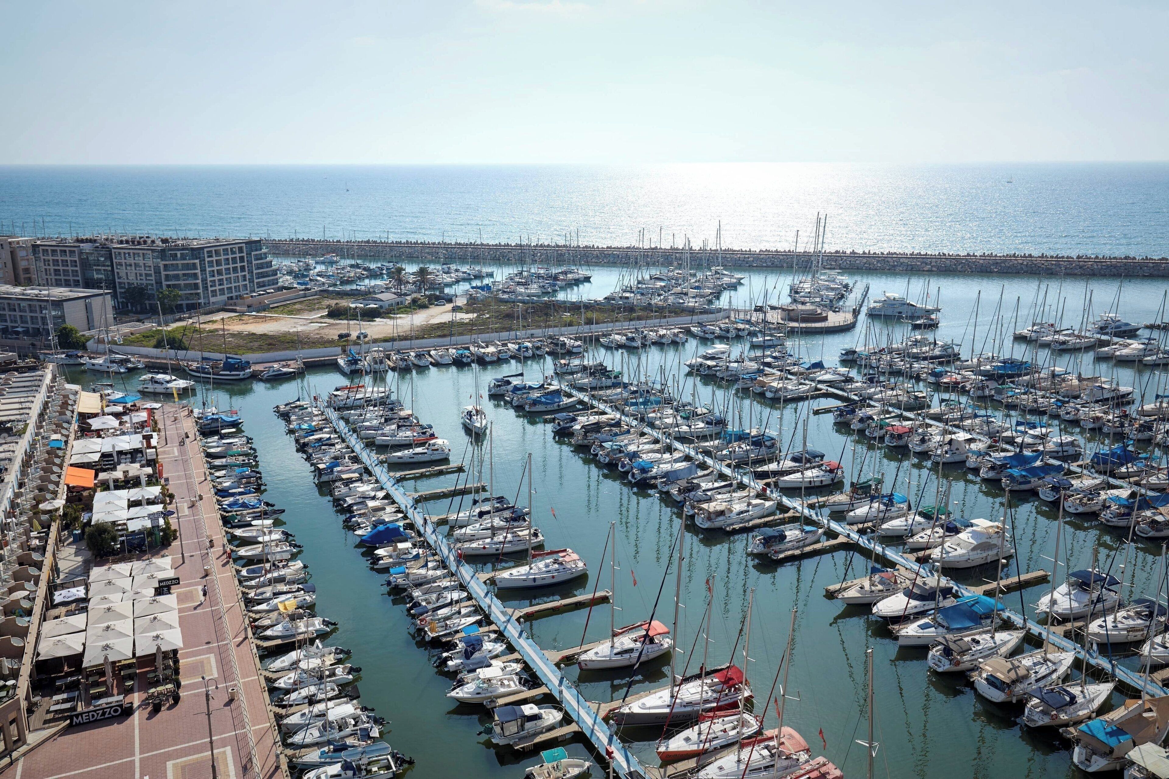 A marina with numerous boats docked in rows, a promenade with outdoor seating, and a view of the sea and distant buildings.