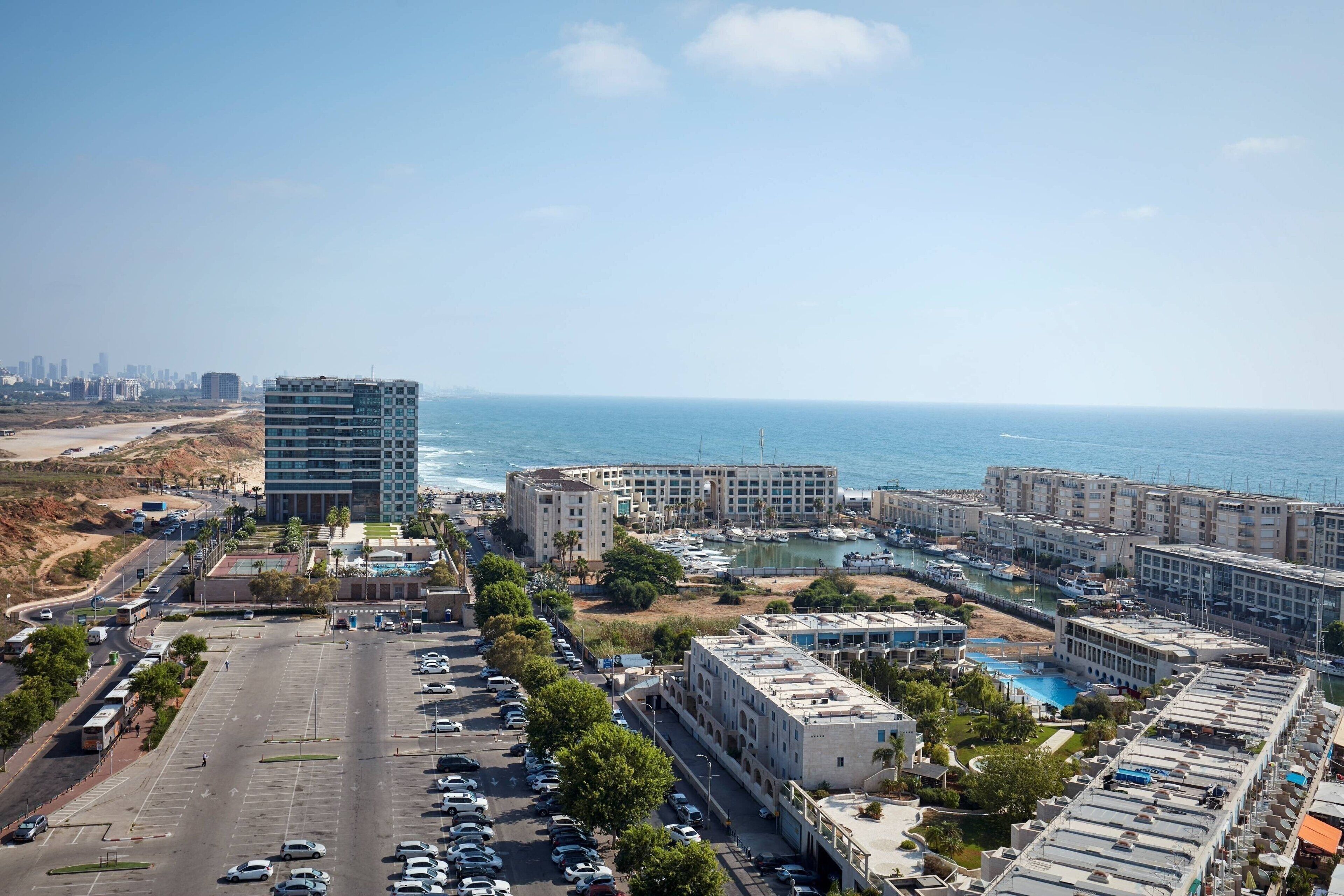 Un paisaje urbano costero con un puerto deportivo, edificios modernos y un estacionamiento.