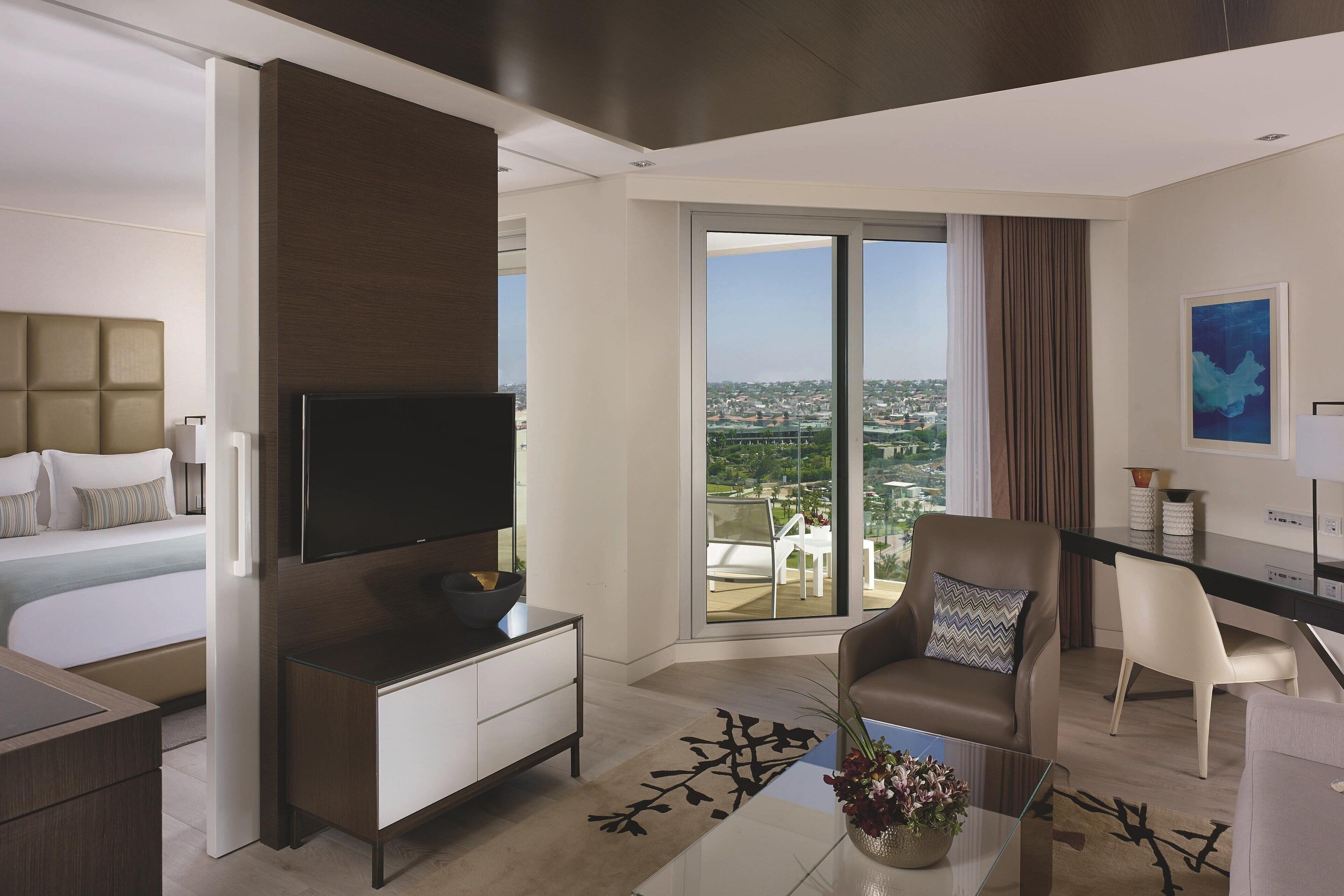 Ein modernes Hotelzimmer mit einem großen Bett, einem Flachbildfernseher, einem Schreibtisch mit Stuhl und einem Balkon mit Blick auf die Stadt.