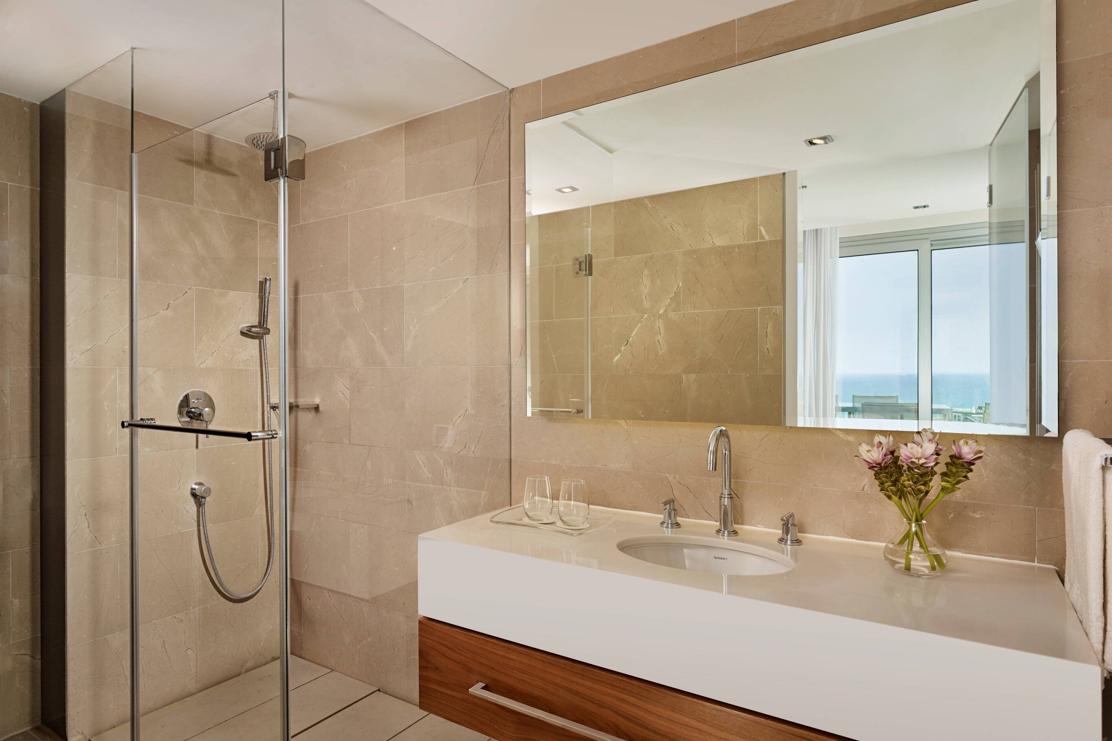 Un baño moderno con cabina de ducha de vidrio, un lavabo blanco y un espejo grande que refleja vistas al exterior.
