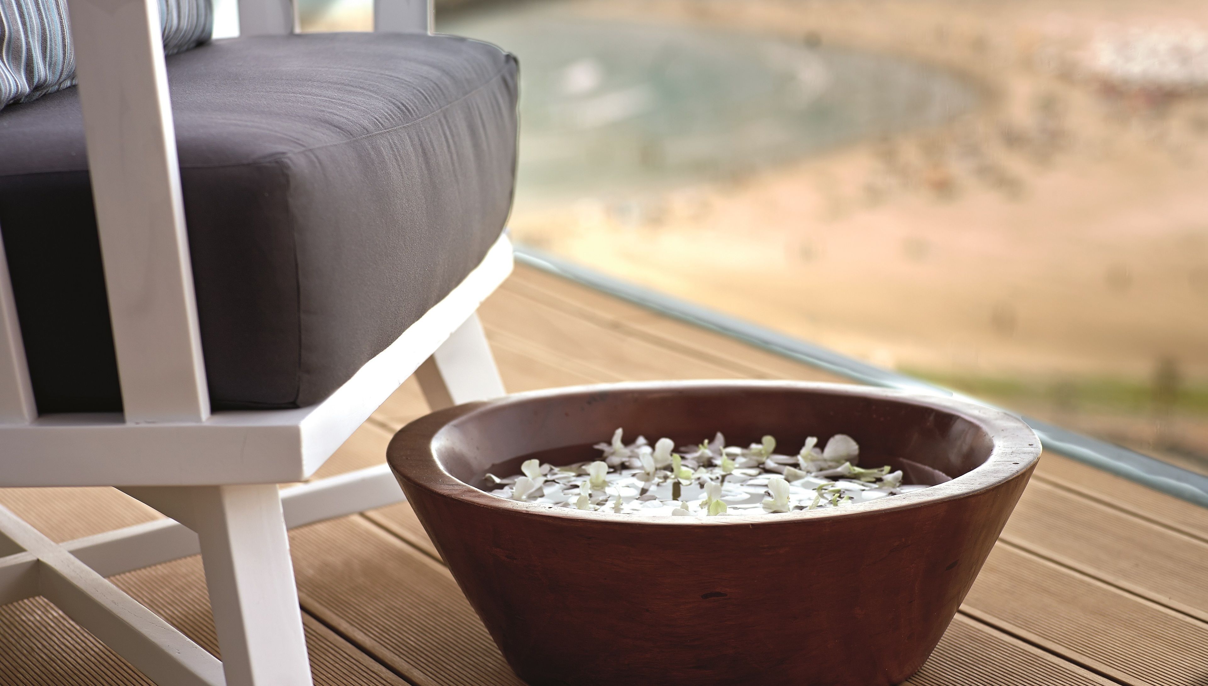 Un saladier en bois orné de fleurs blanches, placé sur une terrasse en bois, avec un canapé gris et une vue sur la plage.