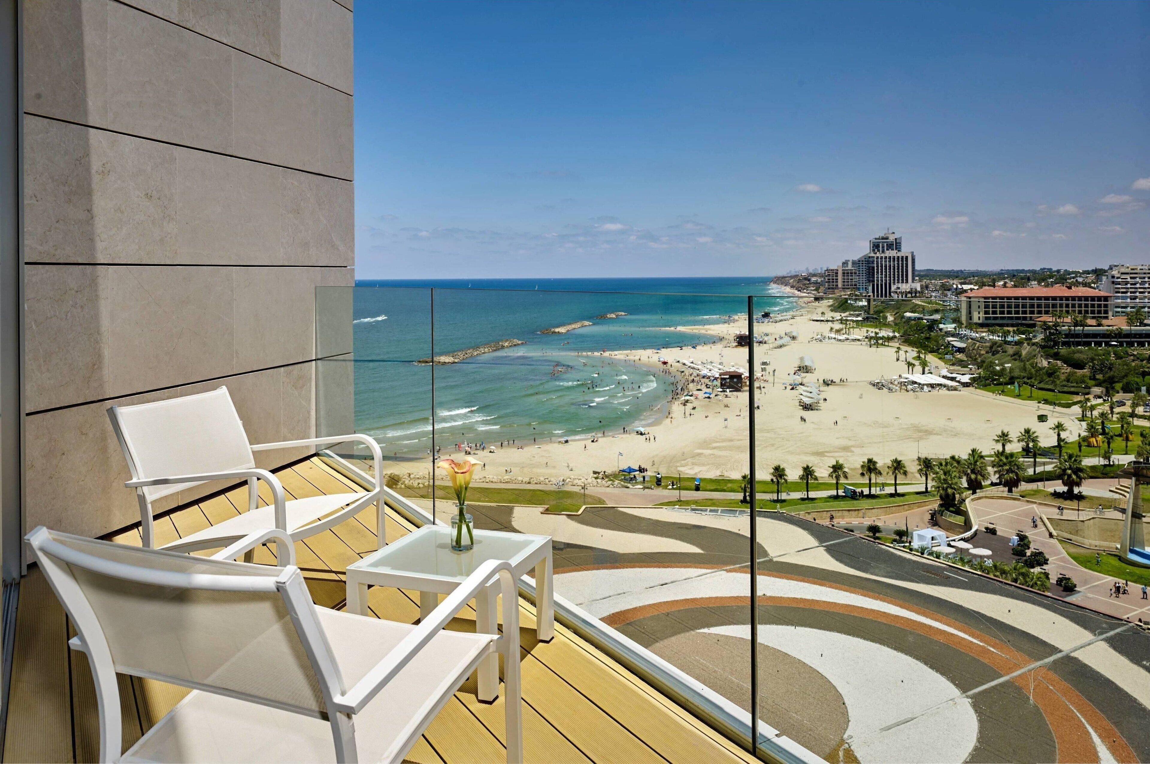 Un balcón con sillas blancas y un jarrón de flores, con vista a una playa y una perspectiva despejada del océano, además de un paisaje urbano distante.