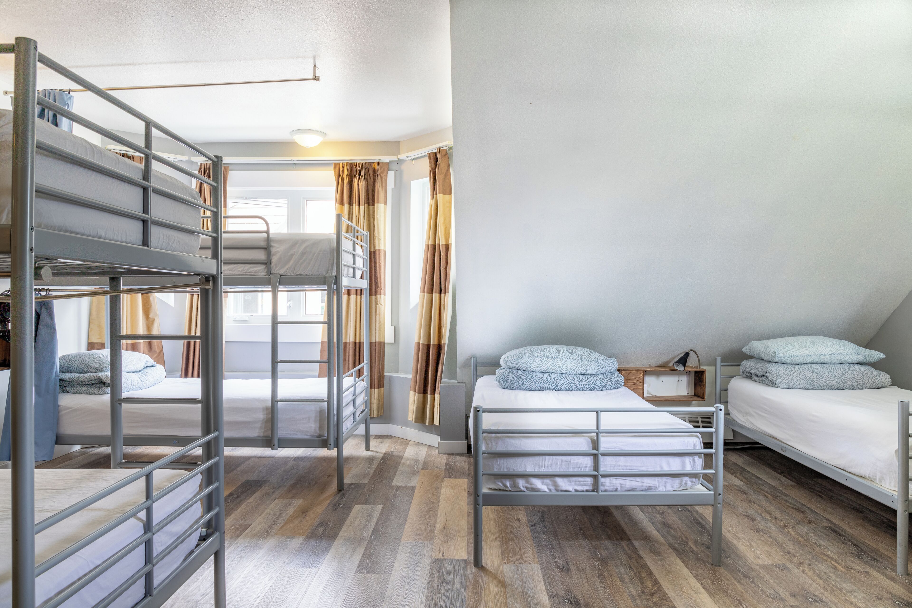 Una habitación tipo dormitorio con literas, suelo de madera, ventana con cortinas y una mesita de noche pequeña.