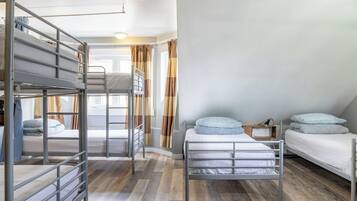 Una habitación tipo dormitorio con literas, suelo de madera, ventana con cortinas y una mesita de noche pequeña.