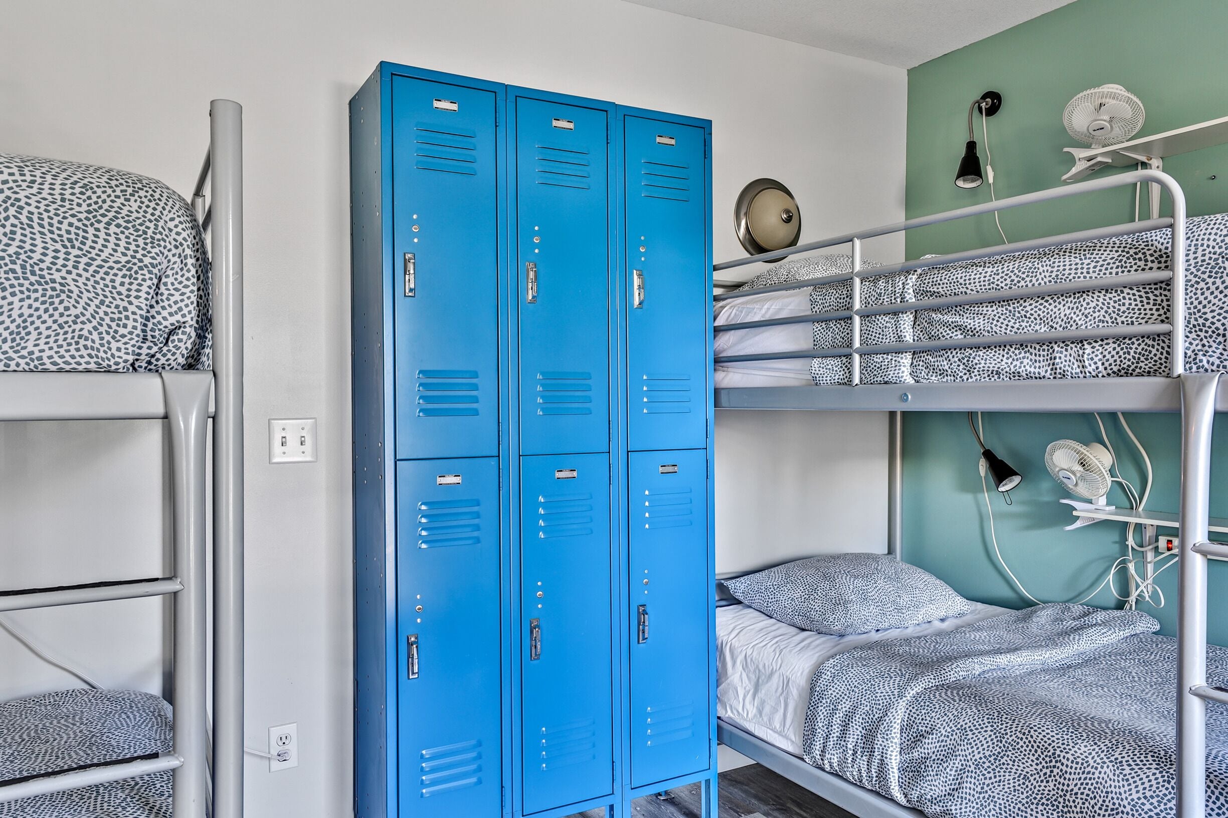 Une chambre avec des lits superposés, un ensemble de casiers bleus et un ventilateur fixé au mur.