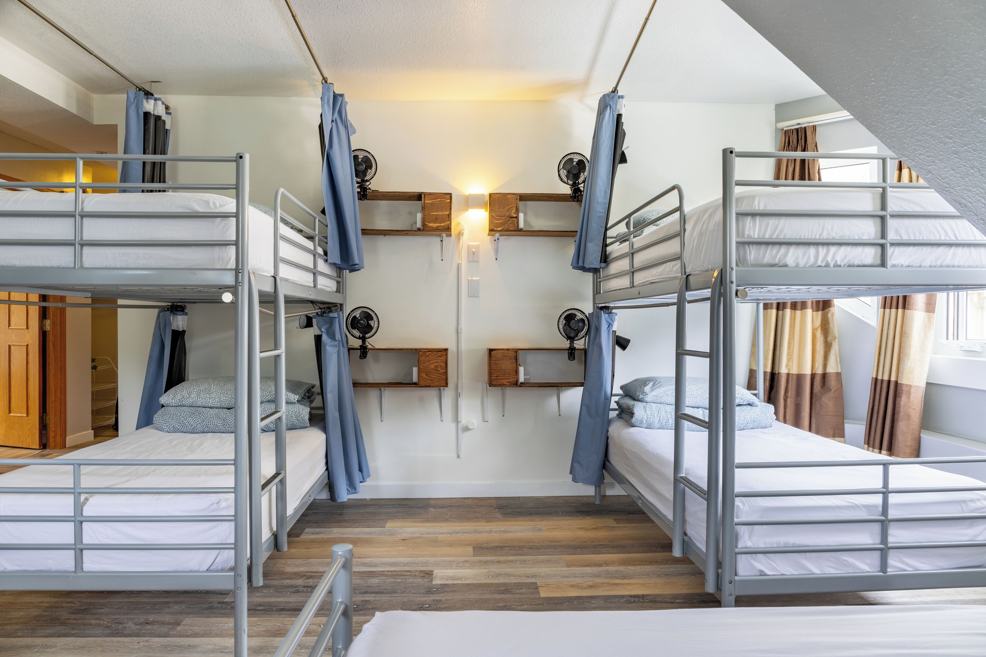 Un dormitorio con literas, suelo de madera, una luz en la pared y una ventana con cortinas.