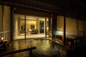 Un área de aguas termales con terraza de madera y cuenca de piedra, conectada a una habitación con sofá y lámpara.