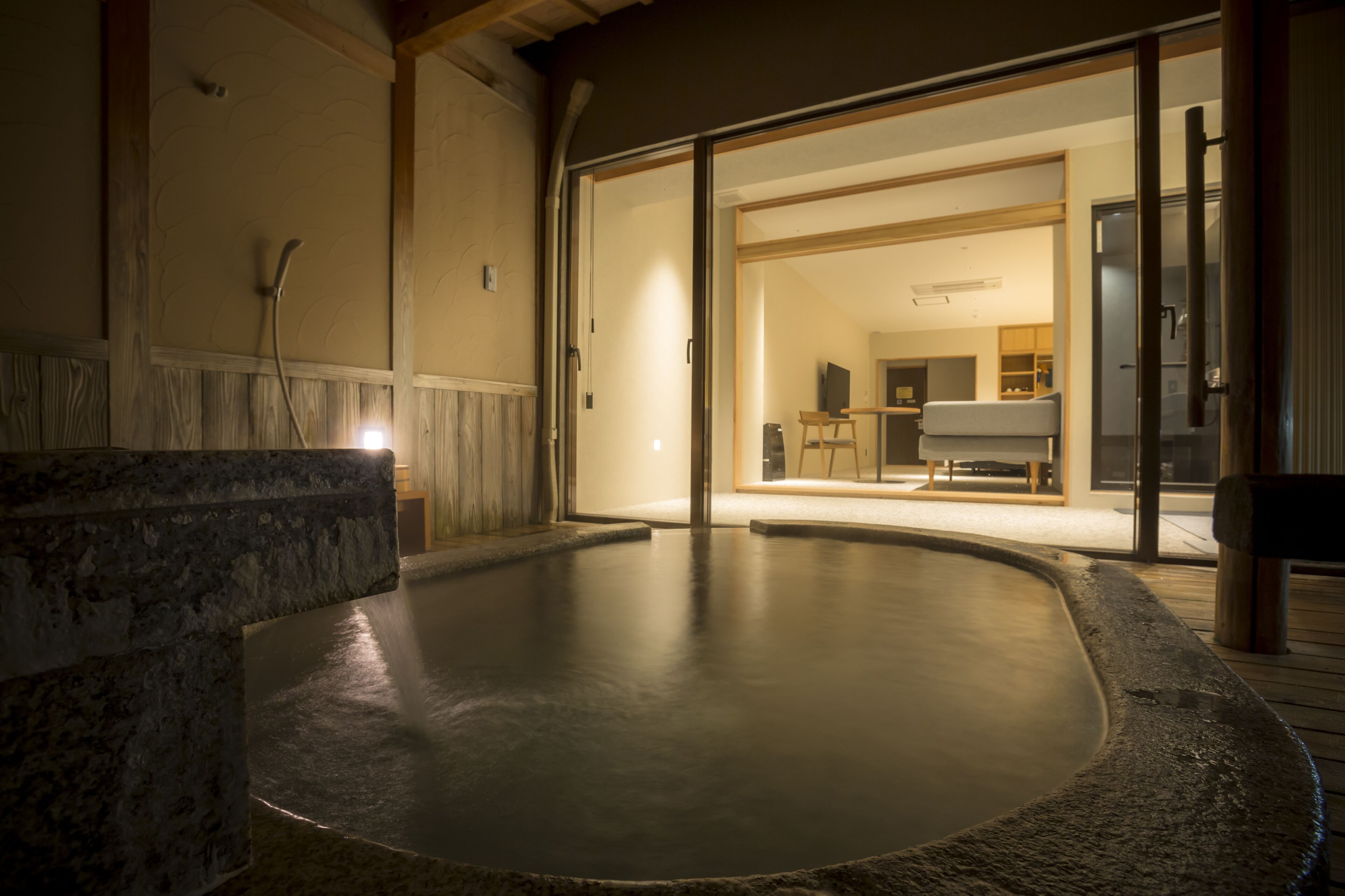 A hot spring bath area with a stone edge, wooden flooring, and a view into a room with a table and chairs.
