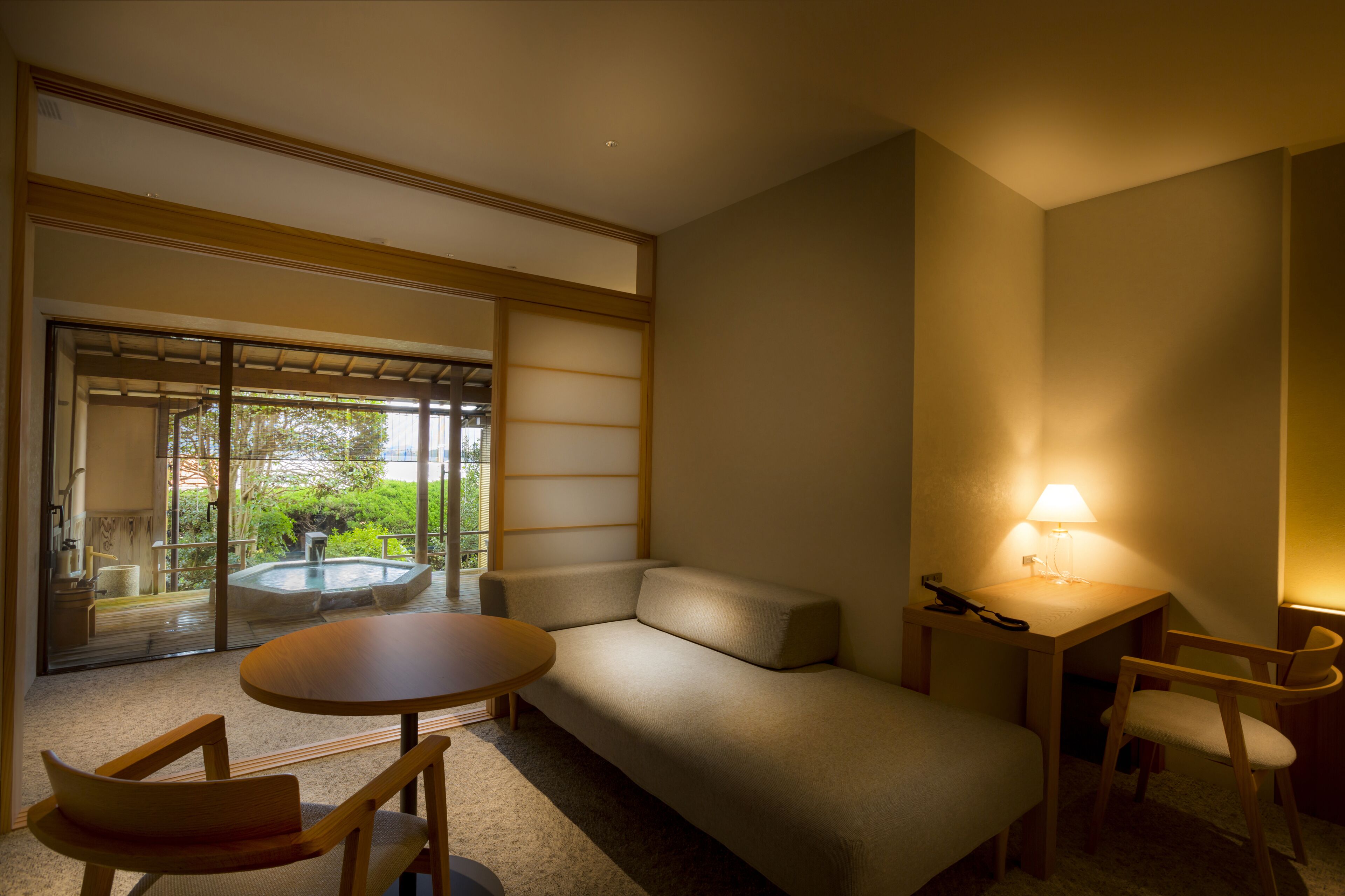 A modern hotel room with a sofa, a small round table, two wooden chairs, a desk with a lamp, and a sliding glass door leading to a balcony with a hot tub.