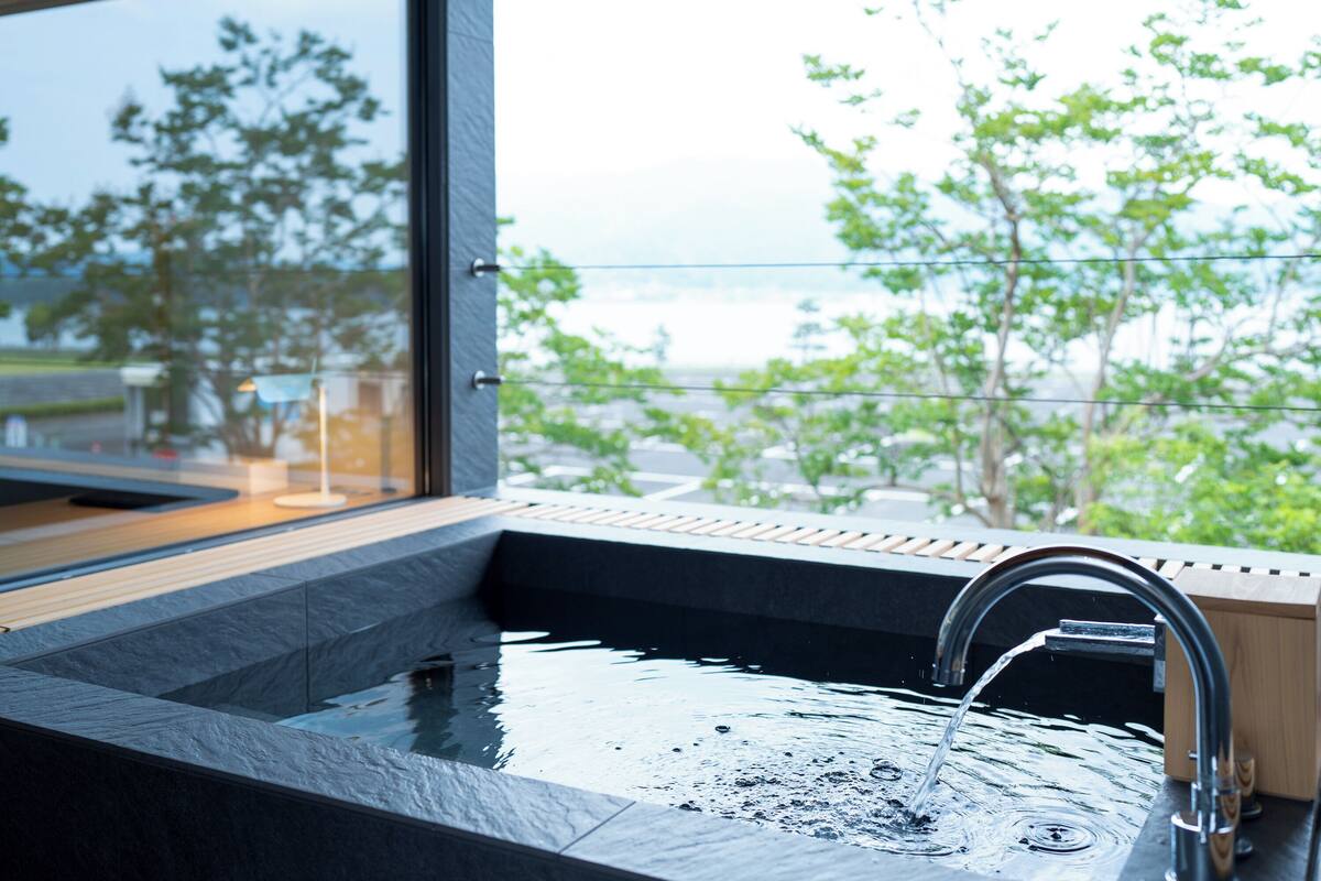 A modern bathroom with a large window, a built-in sink, and a view of greenery and a body of water.