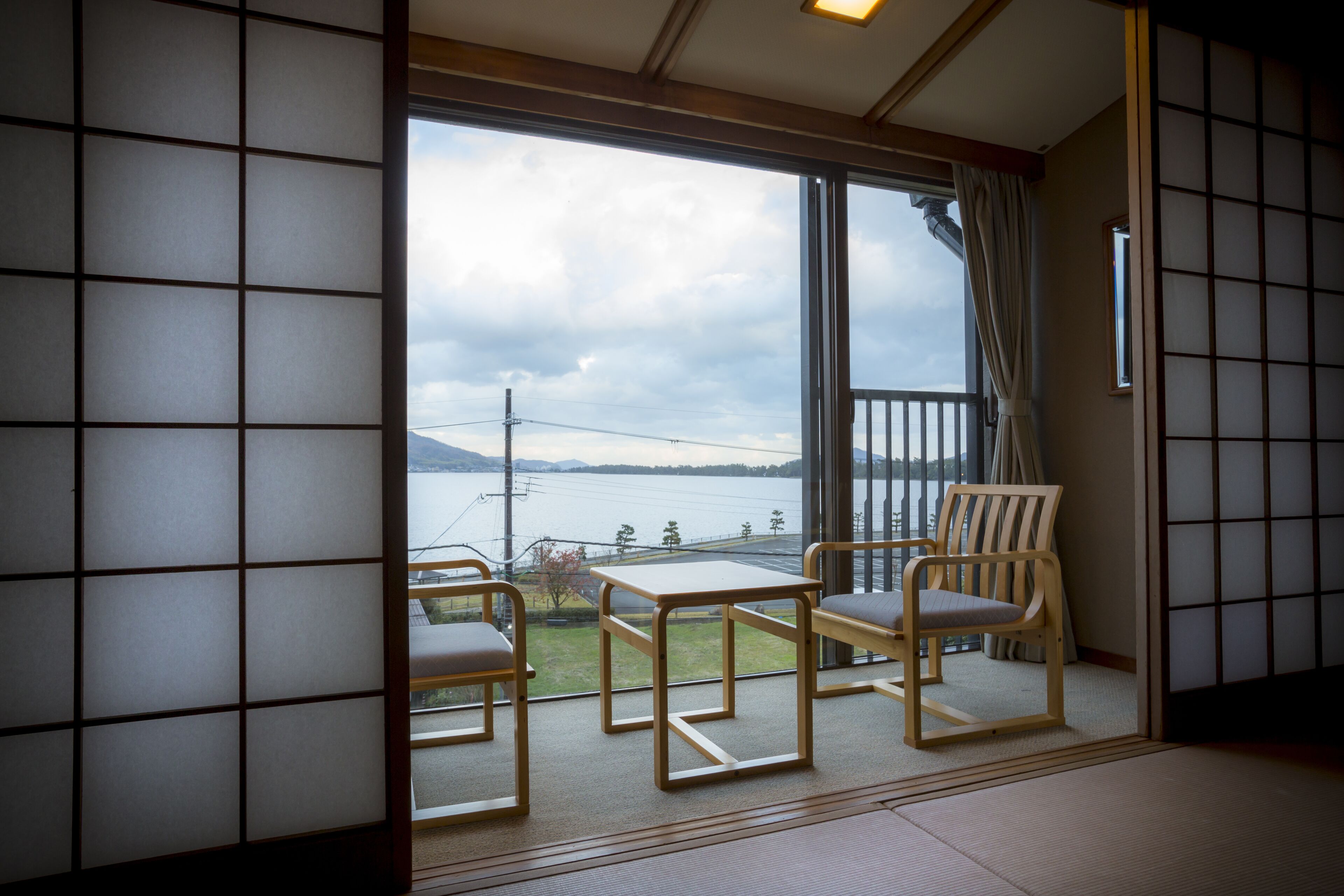 A room with tatami flooring, a sliding glass door leading to a balcony with a view of a body of water and mountains. There is a wooden table and two chairs.