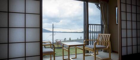 A room with tatami flooring, a sliding glass door leading to a balcony with a view of a body of water and mountains. There is a wooden table and two chairs.