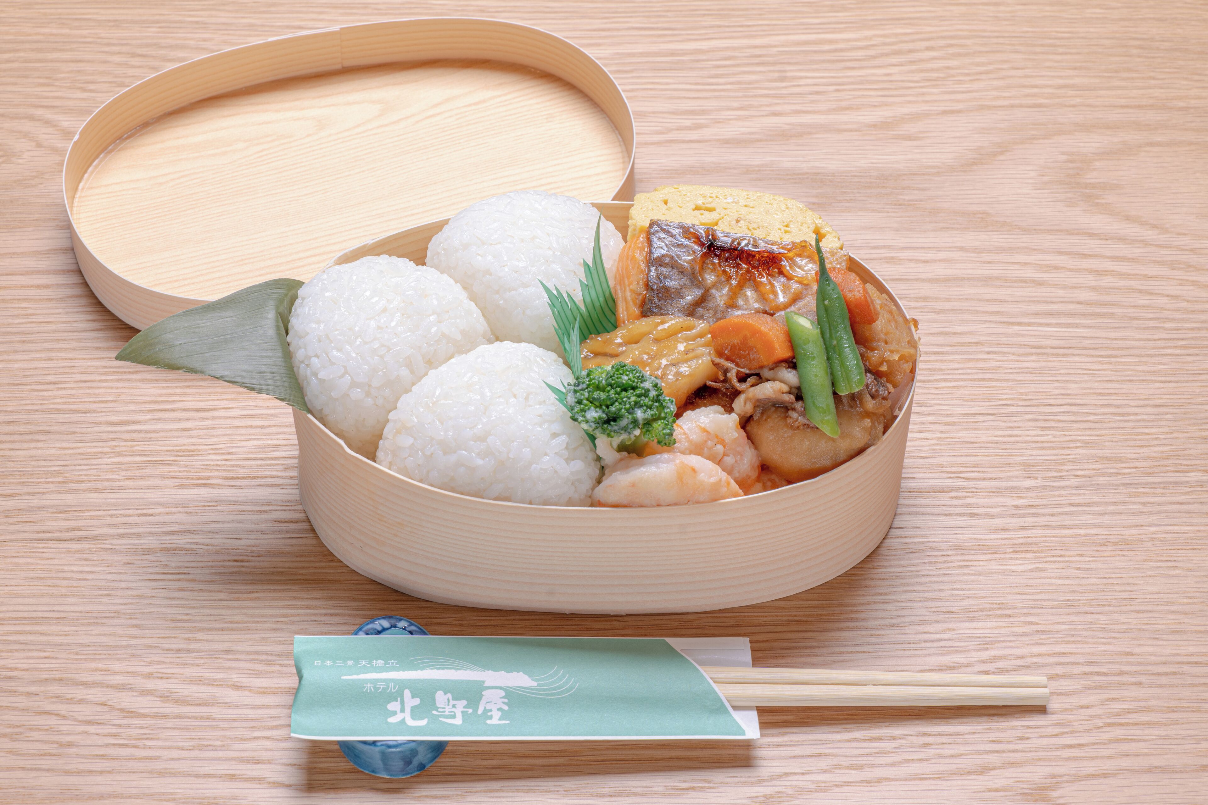 Una caja bento con arroz, pescado, verduras y un trozo de tofu sobre una mesa de madera.