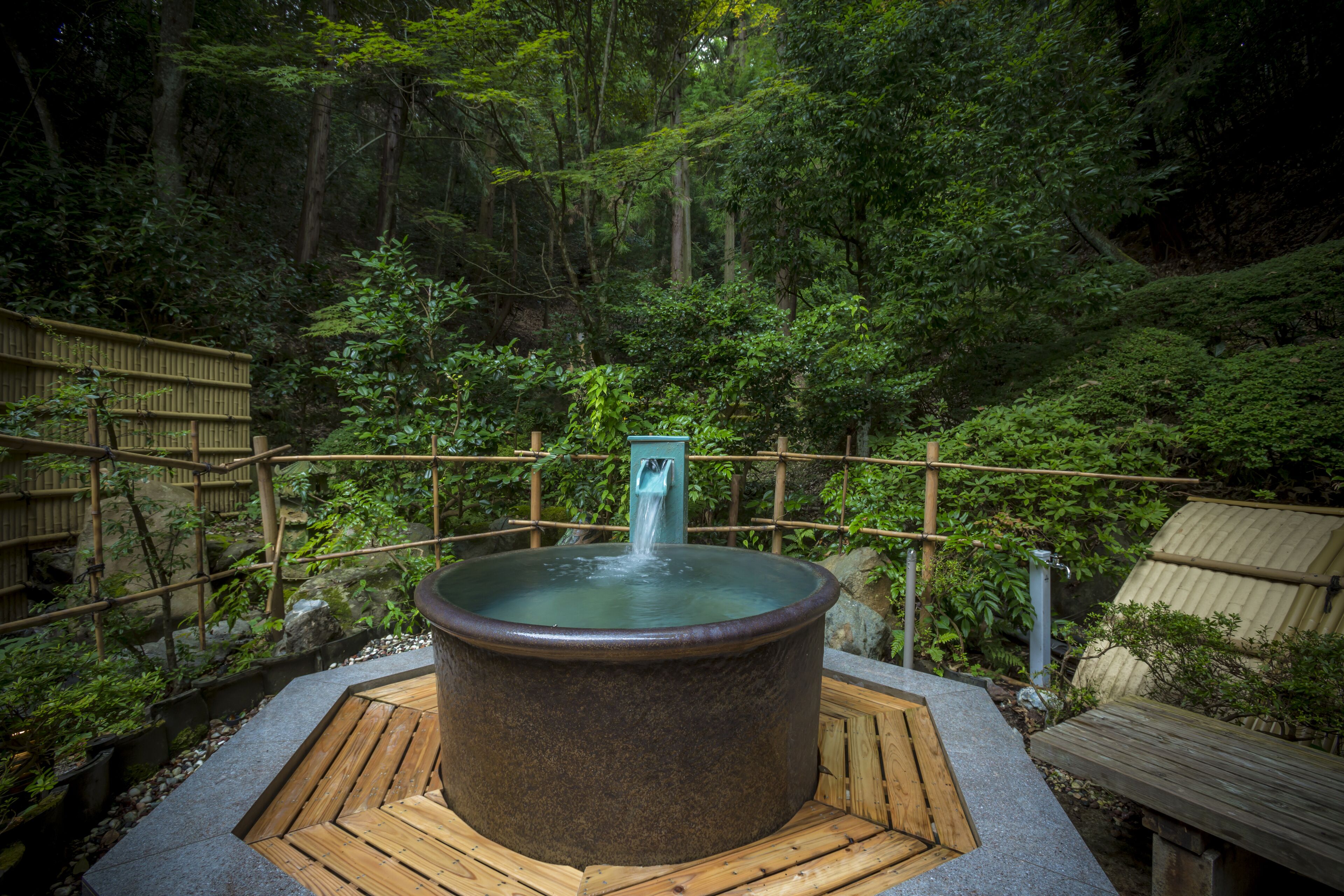 A hot spring bath surrounded by a wooden deck and bamboo fence, set in a lush forested area.