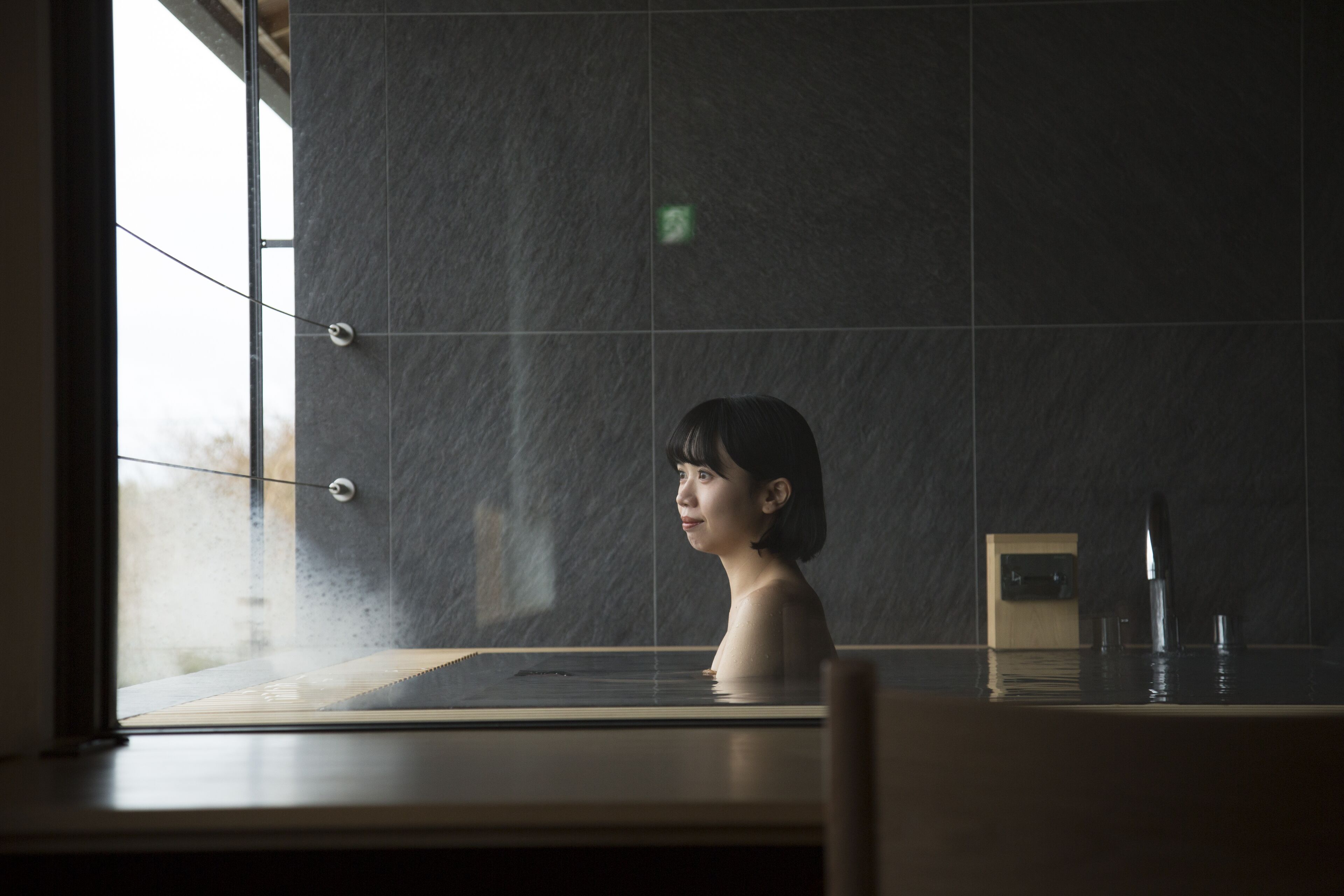 A person in a bathtub with a window and a wall-mounted faucet.