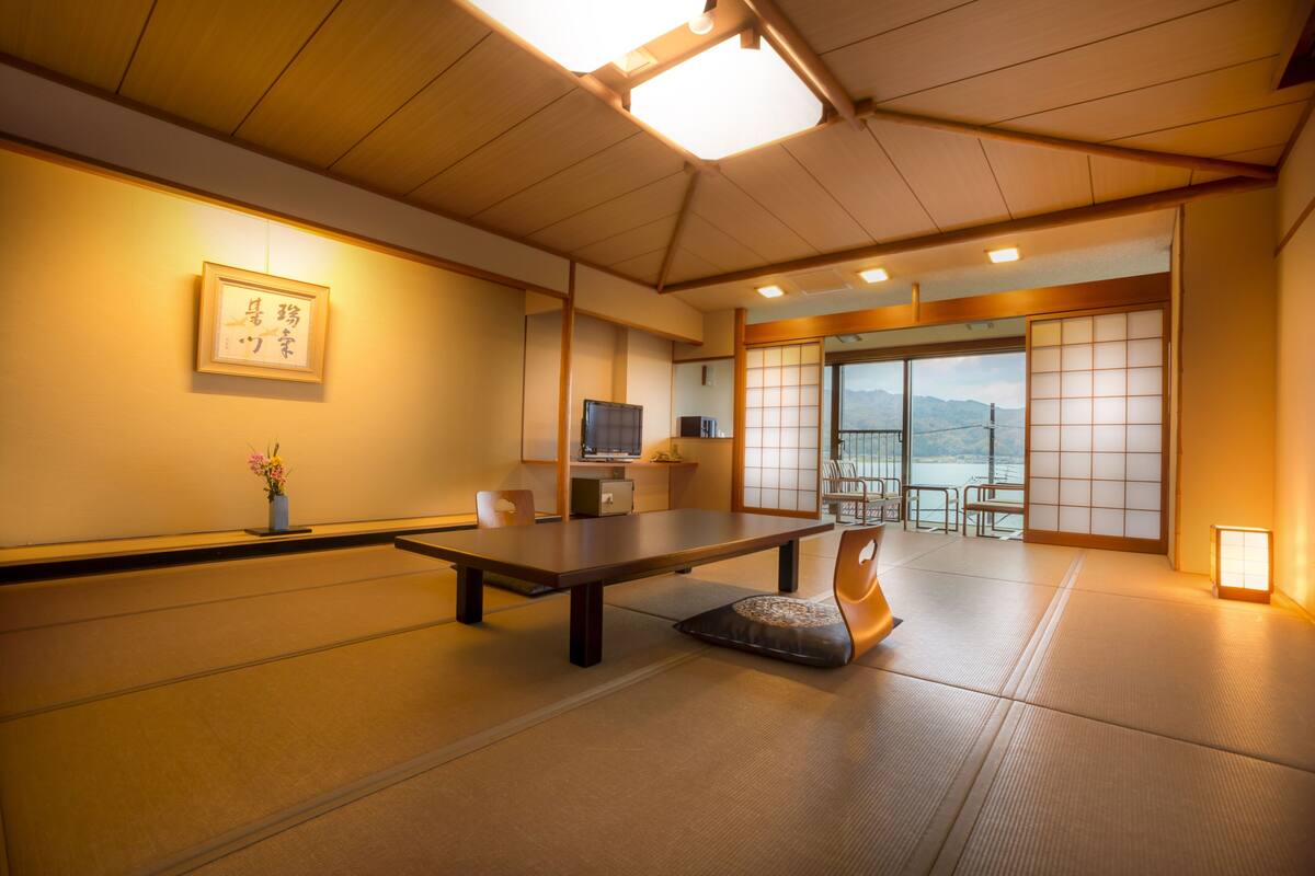 A traditional Japanese room with tatami flooring, a low table, a chair, a TV, and a sliding door leading to a balcony with a view of mountains.