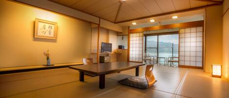 Una habitación japonesa tradicional con suelo de tatami, una mesa baja, una silla, un televisor y una puerta corrediza que da a un balcón con vistas a las montañas.