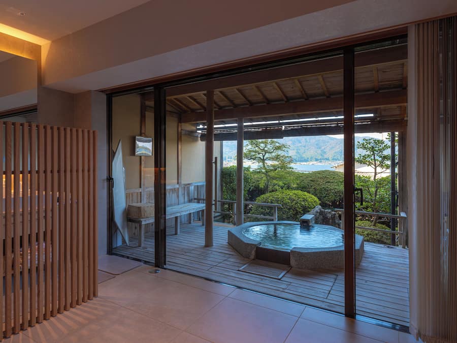Un interior moderno con un divisor de vidrio corredera que da a una terraza exterior con jacuzzi y zona de estar, con vistas a un paisaje pintoresco.