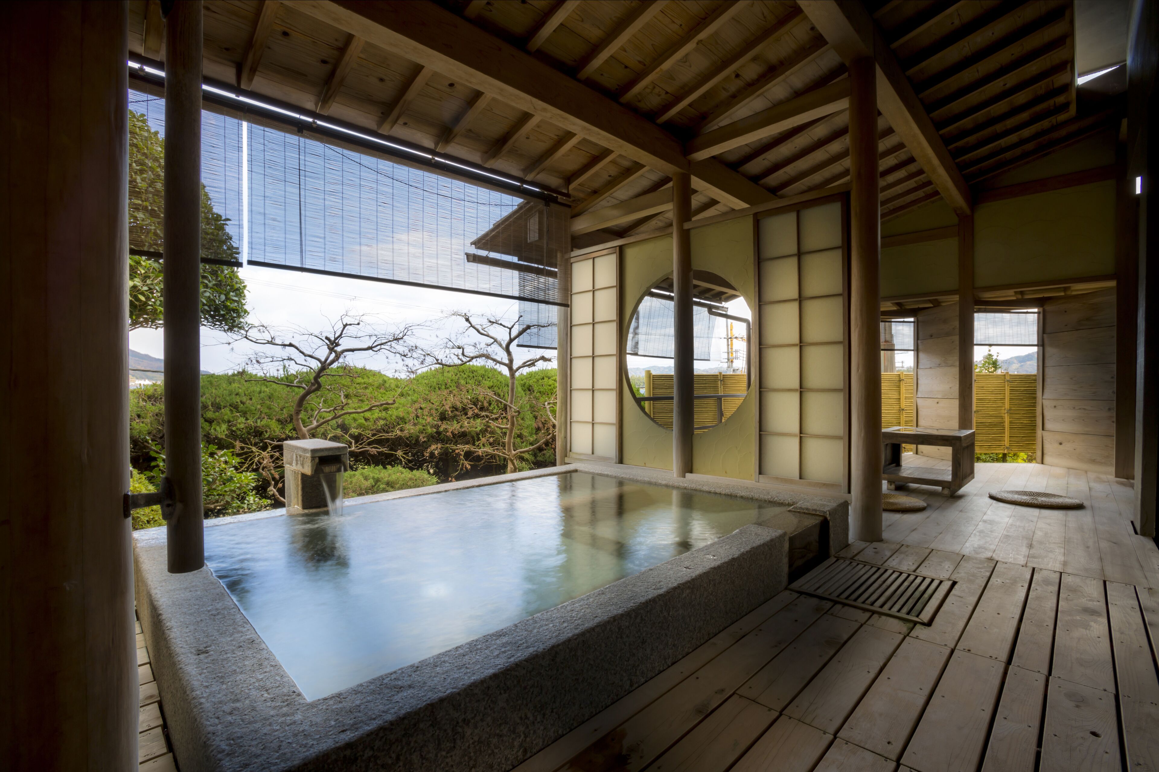 Un onsen japonais traditionnel doté d’une source d’eau chaude naturelle entourée de pierres, d’une terrasse en bois, d’un sol recouvert de tatamis et offrant une vue sur la nature environnante.