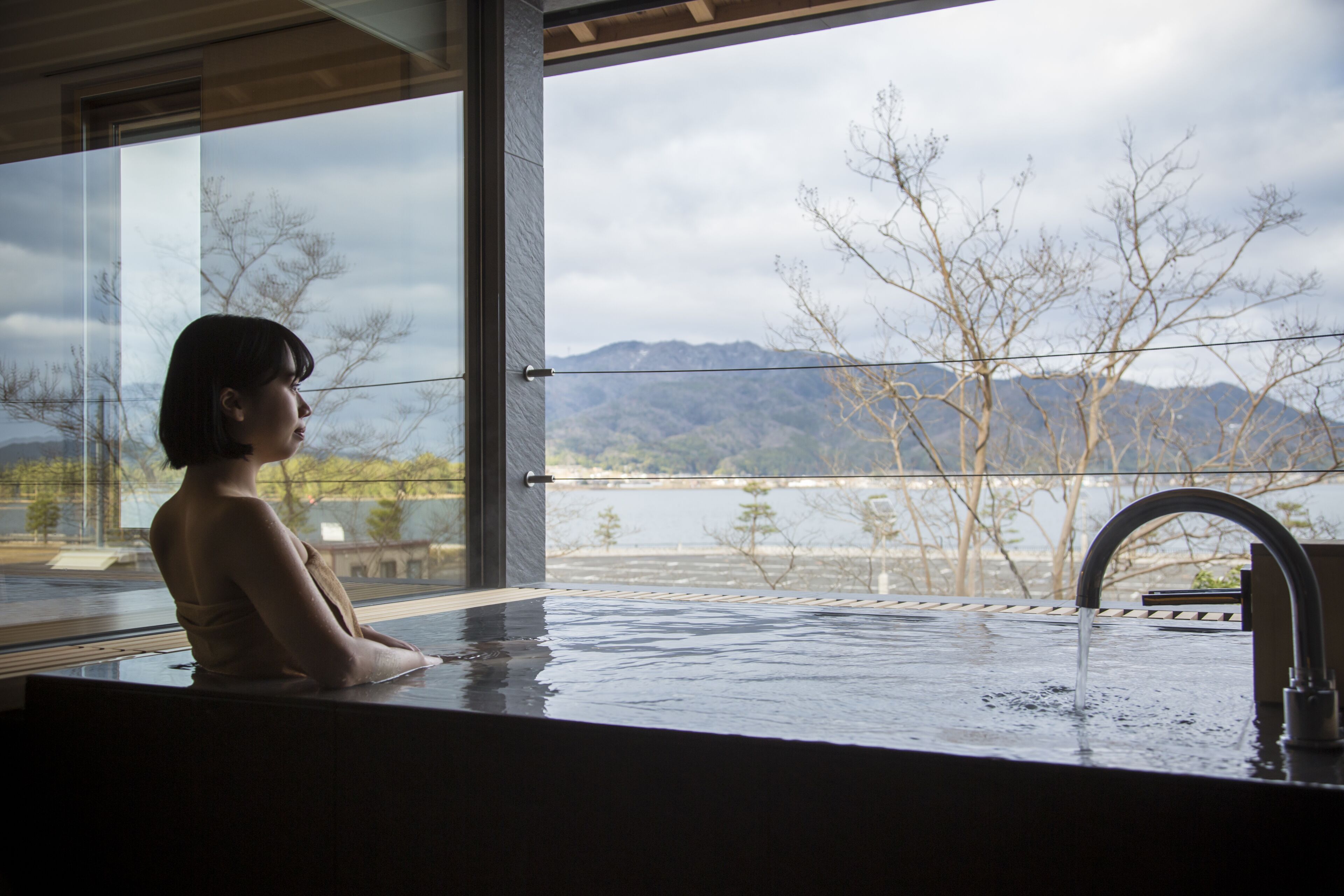 Una persona sentada en una bañera con vistas a montañas y árboles a través de un ventanal amplio.