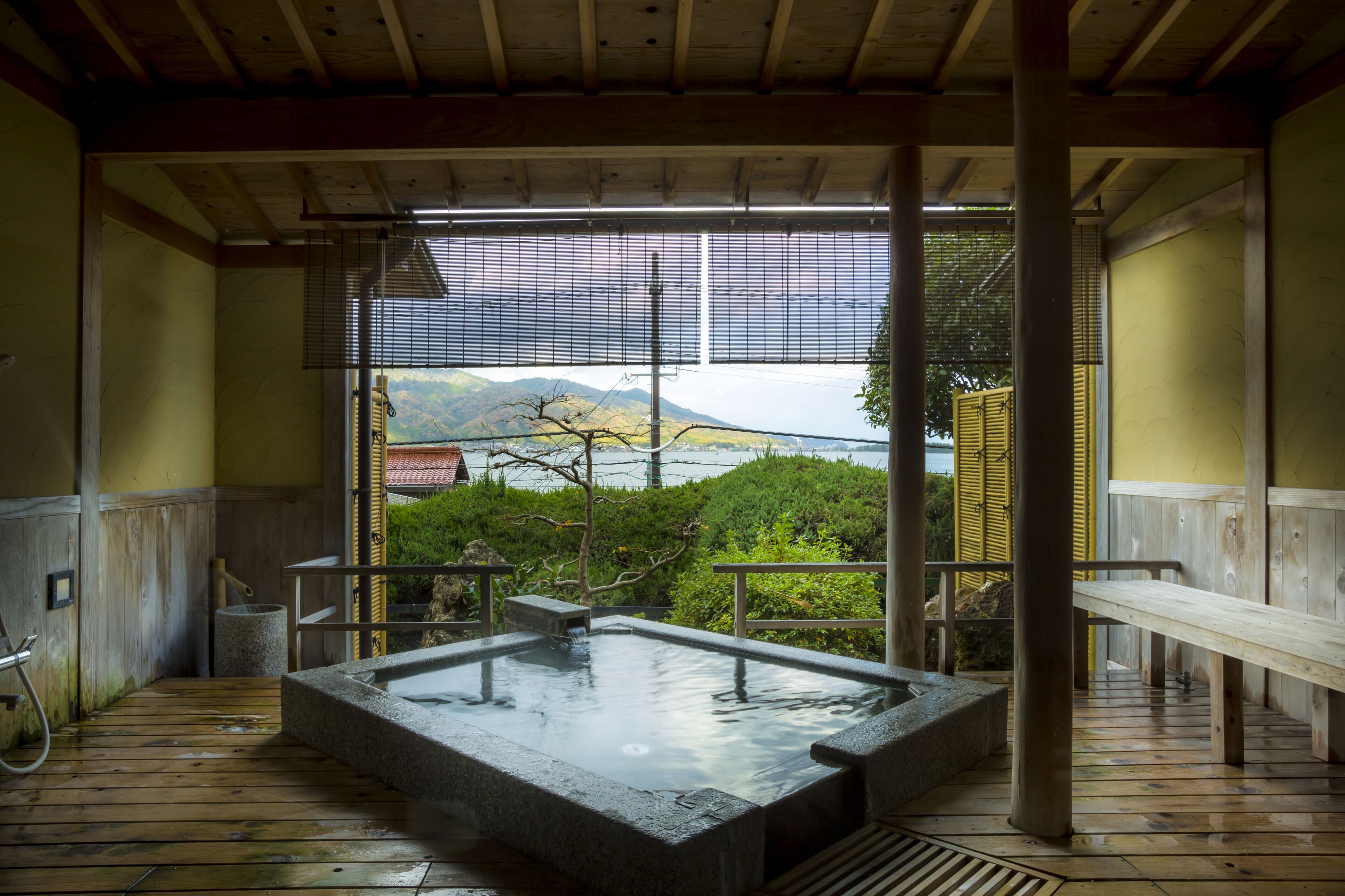Un onsen japonais traditionnel doté d’une terrasse en bois, d’une source thermale en pierre et offrant une vue sur les montagnes et l’eau.