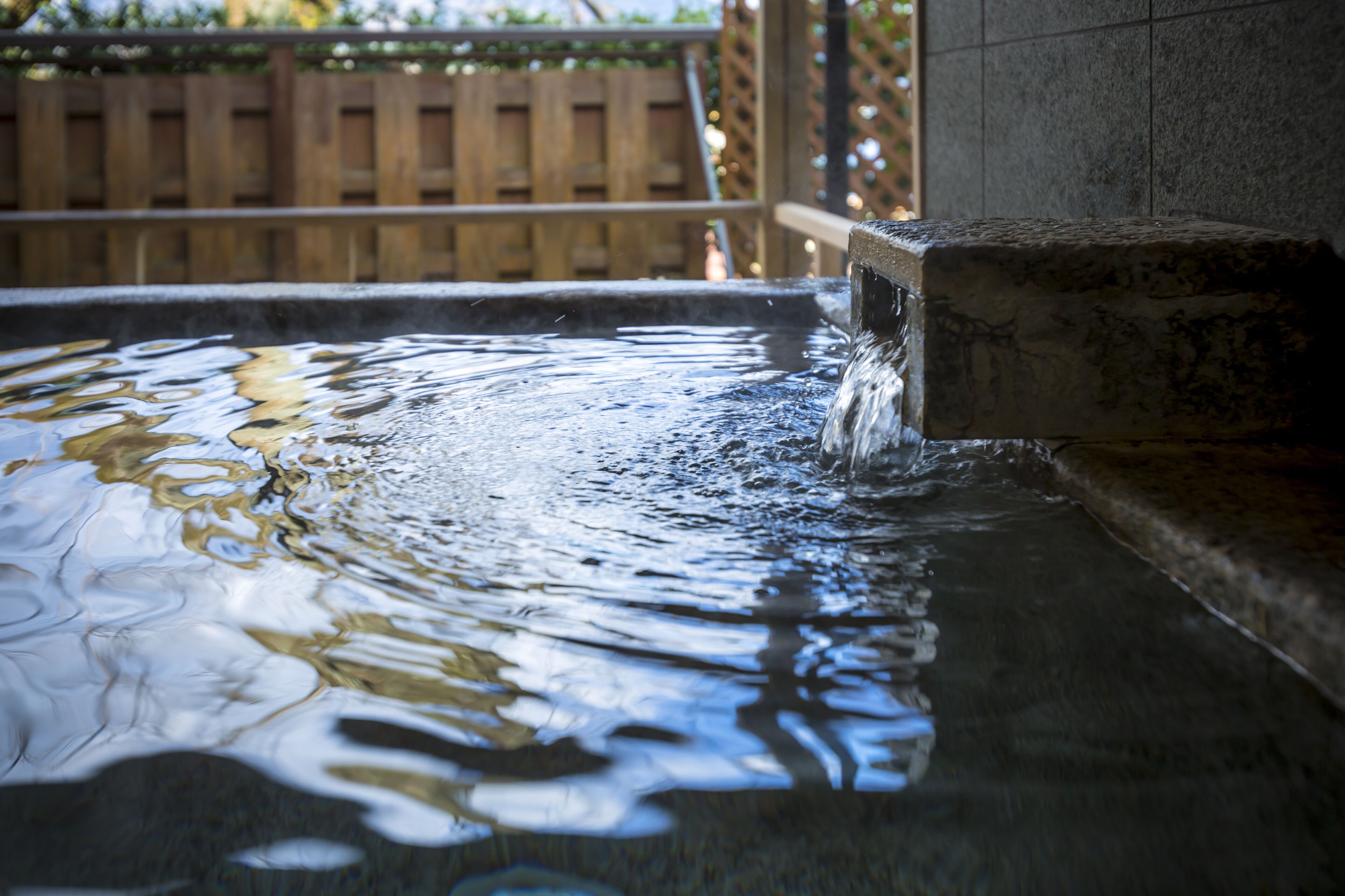 A hot tub with water flowing into it, surrounded by a wooden fence and a stone wall.