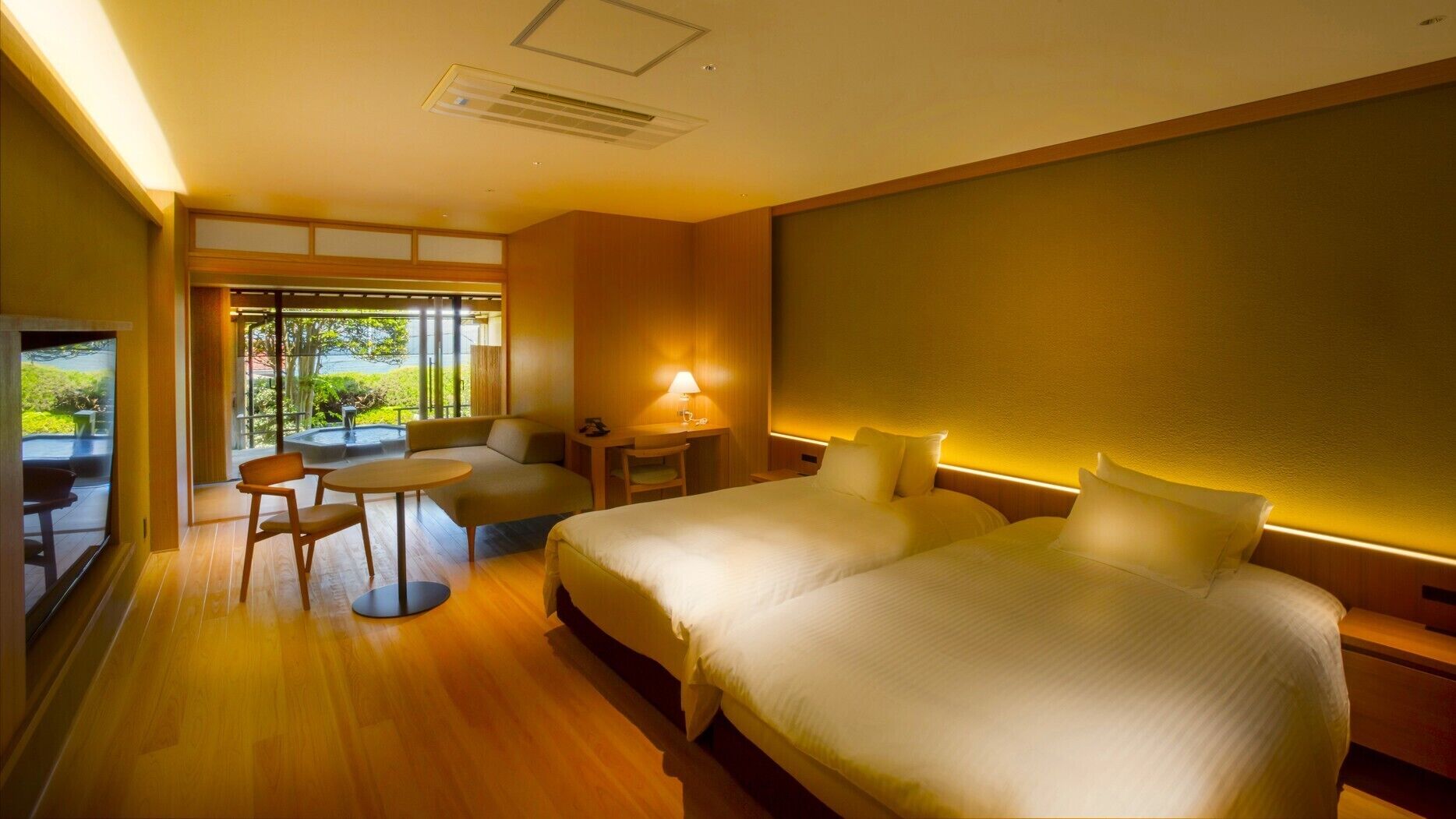 A hotel room with a large bed, a desk, a chair, and a small table. There is a window with a view of greenery.