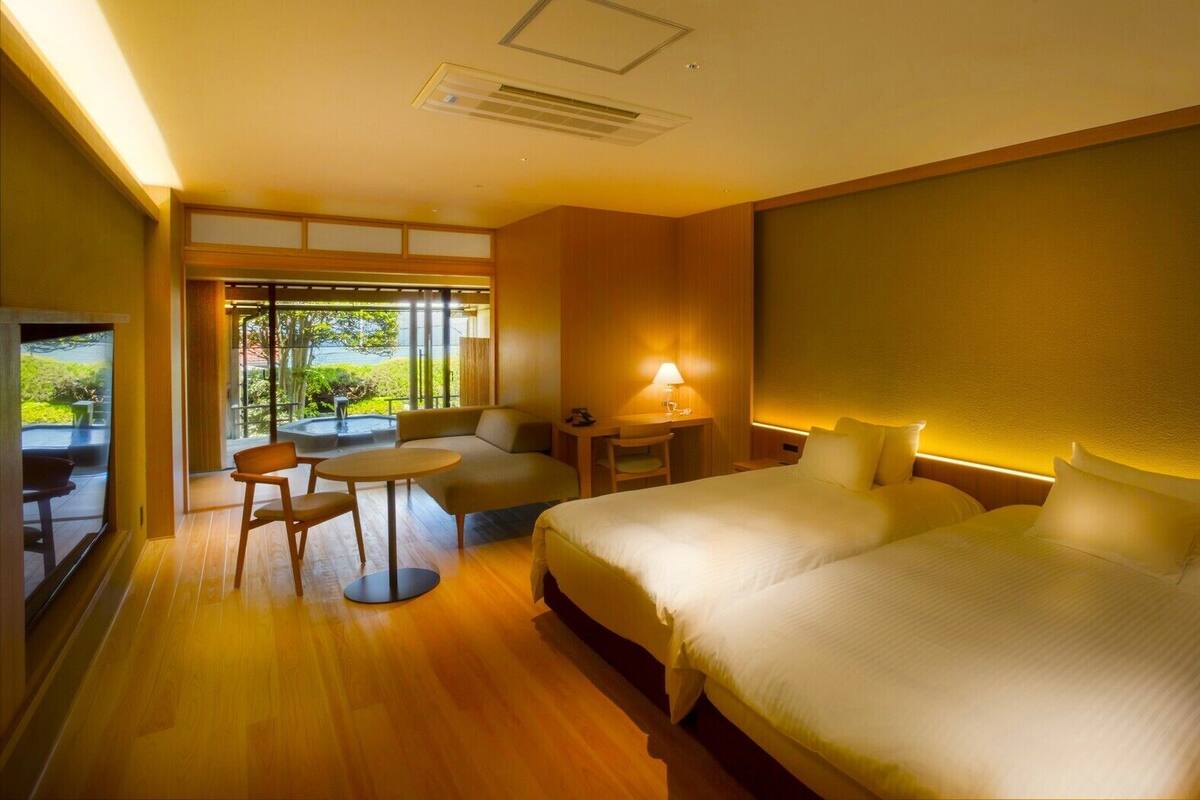 A hotel room with a large bed, a desk, a chair, and a small table. There is a window with a view of greenery.