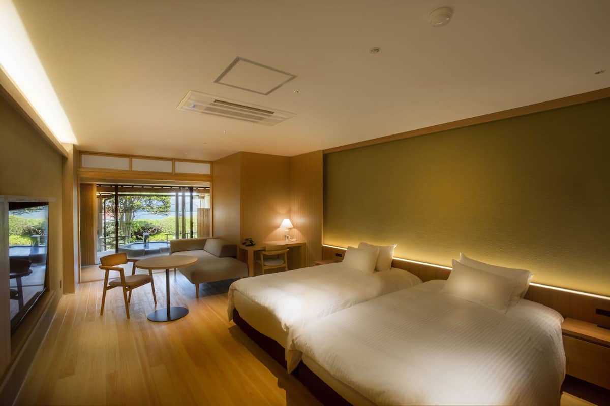 A hotel room with a large bed, a desk, a chair, and a small table. There is a window with a view of greenery outside.