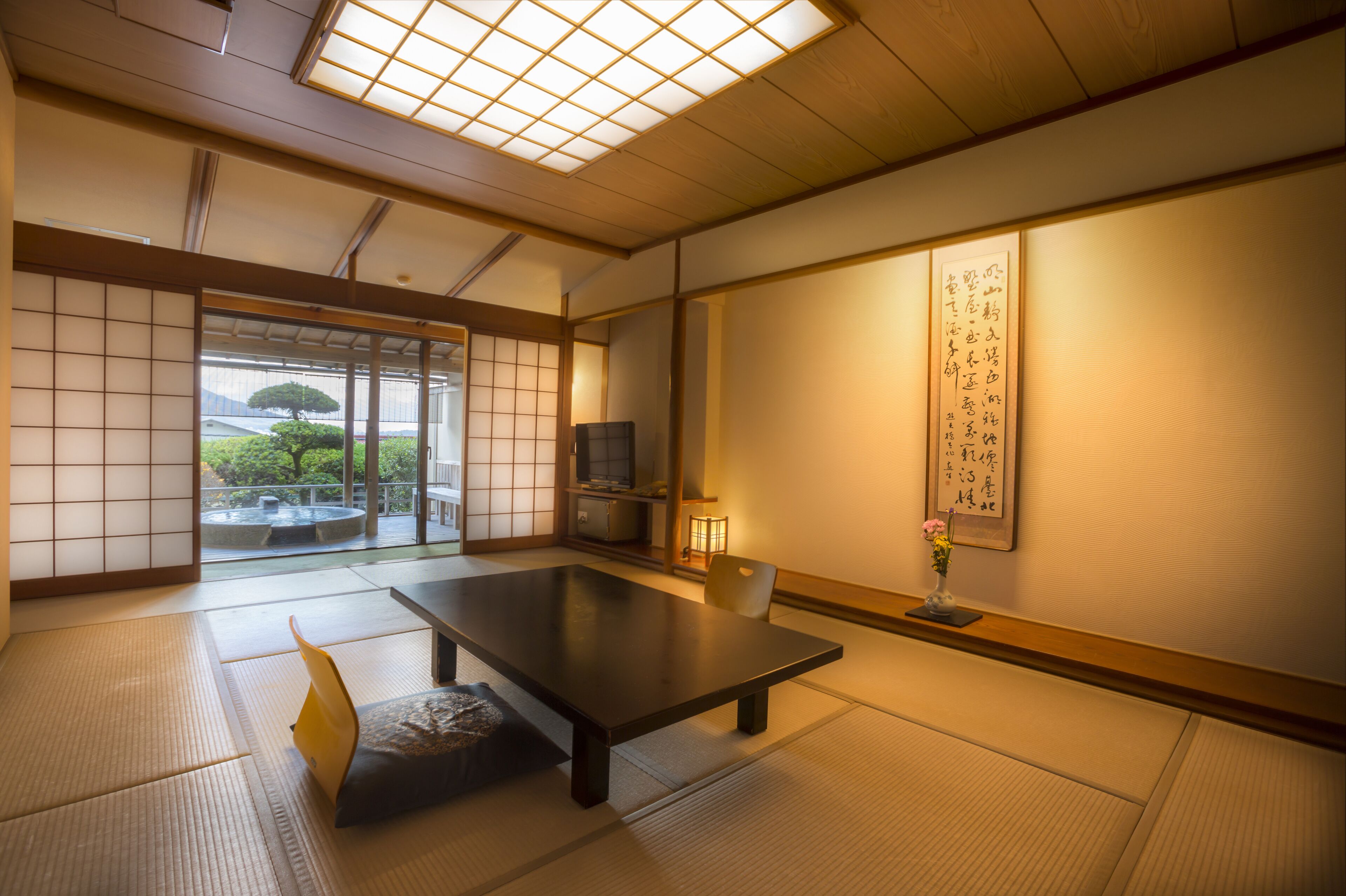 A traditional Japanese room with tatami flooring, a low table, sliding doors, and a view of greenery outside.